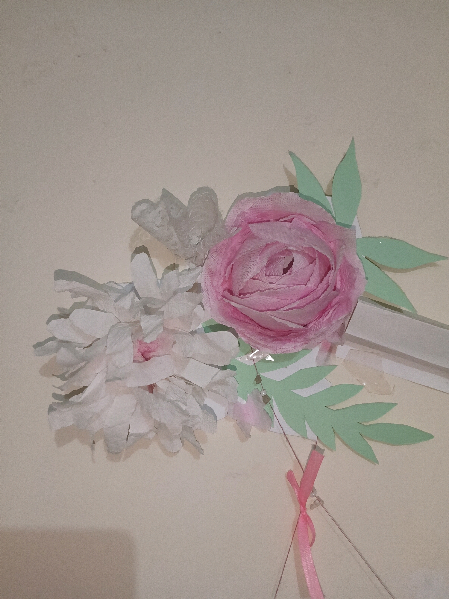A close-up, low-angle, indoor photograph displays a handmade floral arrangement against a neutral, textured wall. The arrangement features a large, detailed pink rose with ruffled petals on the right, and a cluster of white, crepe-paper flowers with hints of pink on the left. Several light green paper leaves are artfully placed behind and around the flowers. A thin, light grey string is visible, attached to a small, pink ribbon tied with a bow, which dangles from the lower right portion of the arrangement. A subtle shadow is cast on the wall from the arrangement. The image provides no information regarding the city of Karnal, India, or any specific context beyond the visual elements present.
