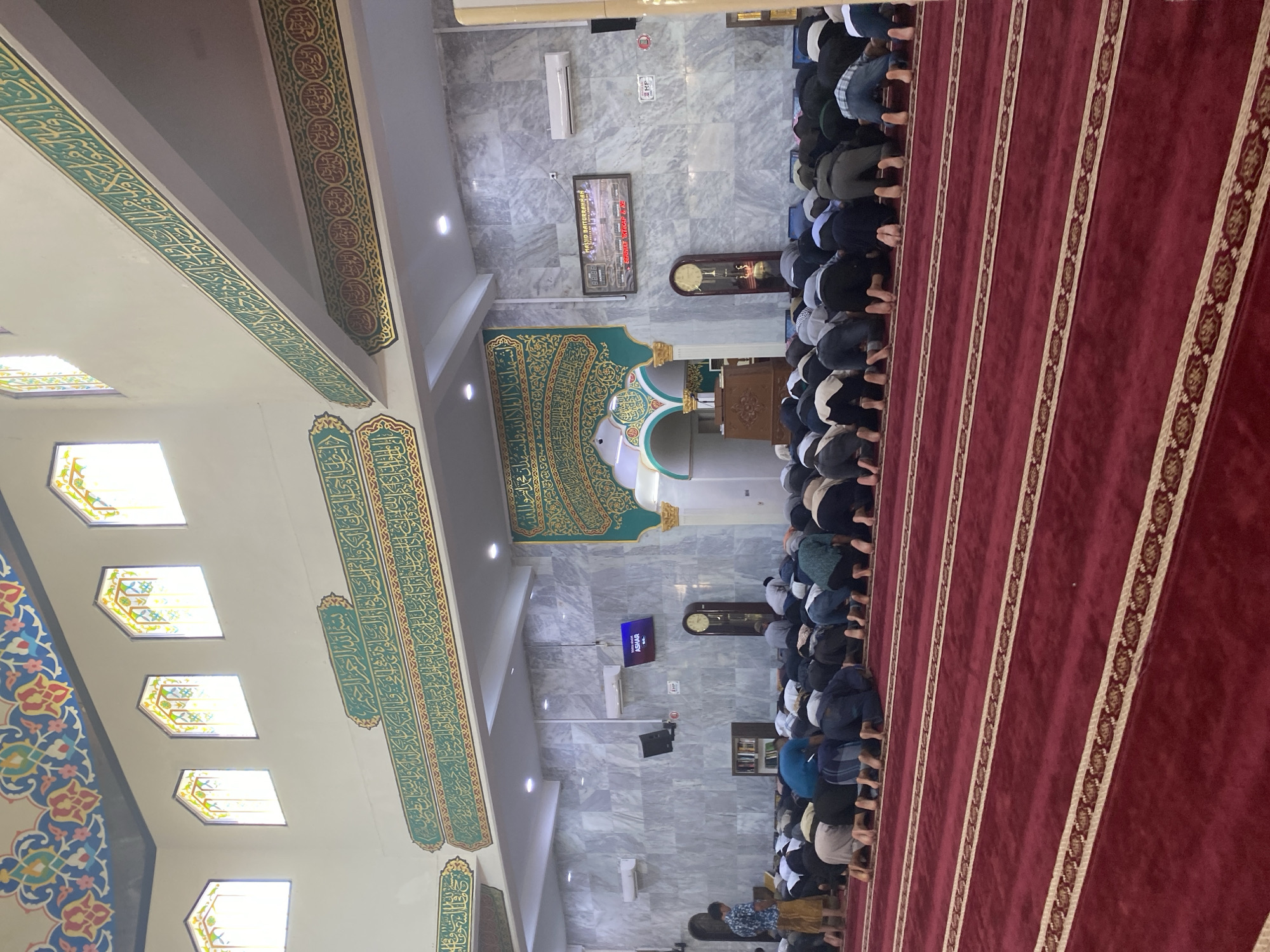 The image shows the interior of the Masjid Baiturrahmah in Lhokseumawe, Indonesia, during prayer. Men are lined up in rows on a red carpet with patterned borders, facing the front of the mosque. Walls and pillars are decorated with Islamic calligraphy. There is a digital display showing the time for prayer. The mosque has marble-tiled walls and multiple windows. A large green and gold decorated arch is visible. Clocks and other notices are on the walls.