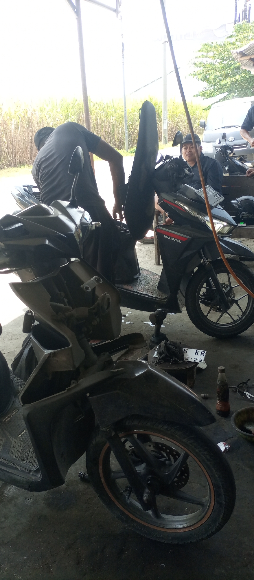 In Sadang, Indonesia, two men are working on a motorcycle under a covered structure. The man on the left, wearing a dark t-shirt and cap, is hunched over and appears to be adjusting something on the motorcycle. To the right, another man in a dark shirt and cap is seated and looking towards the camera. Two motorcycles are visible, one in the foreground, partially disassembled, and another behind it. The environment appears to be an outdoor workshop, with a backdrop of tall grass and trees. There are tools and other motorcycle parts scattered around the ground. A white van is parked in the background.