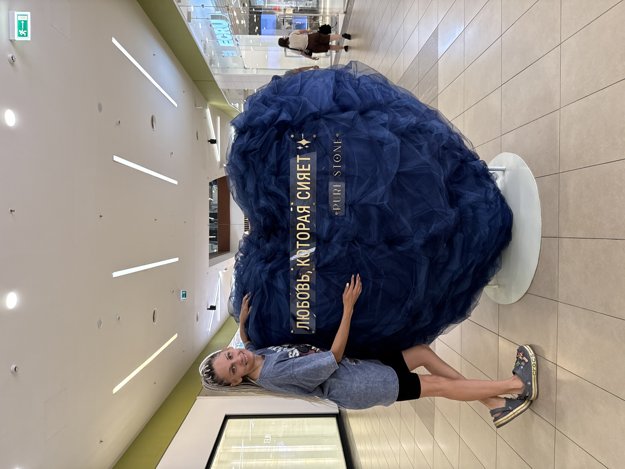 A blonde woman with her hair in braids is posing next to a large, dark blue, heart-shaped object covered in fabric in a shopping mall. The woman is wearing a gray t-shirt, black shorts, and gray Crocs. The heart-shaped object has two signs on it. The top sign reads "ЛЮБОВЬ, КОТОРАЯ СИЯЕТ" which translates to "Love That Shines" in Russian, and the bottom sign reads "PURE STONE." The shopping mall has a tiled floor, bright lighting from overhead fixtures, and storefronts in the background. An exit sign with a green running man is visible on the ceiling. In the background, another person is walking away from the camera. The location is identified as the city of Arkhangelsk, Russia.