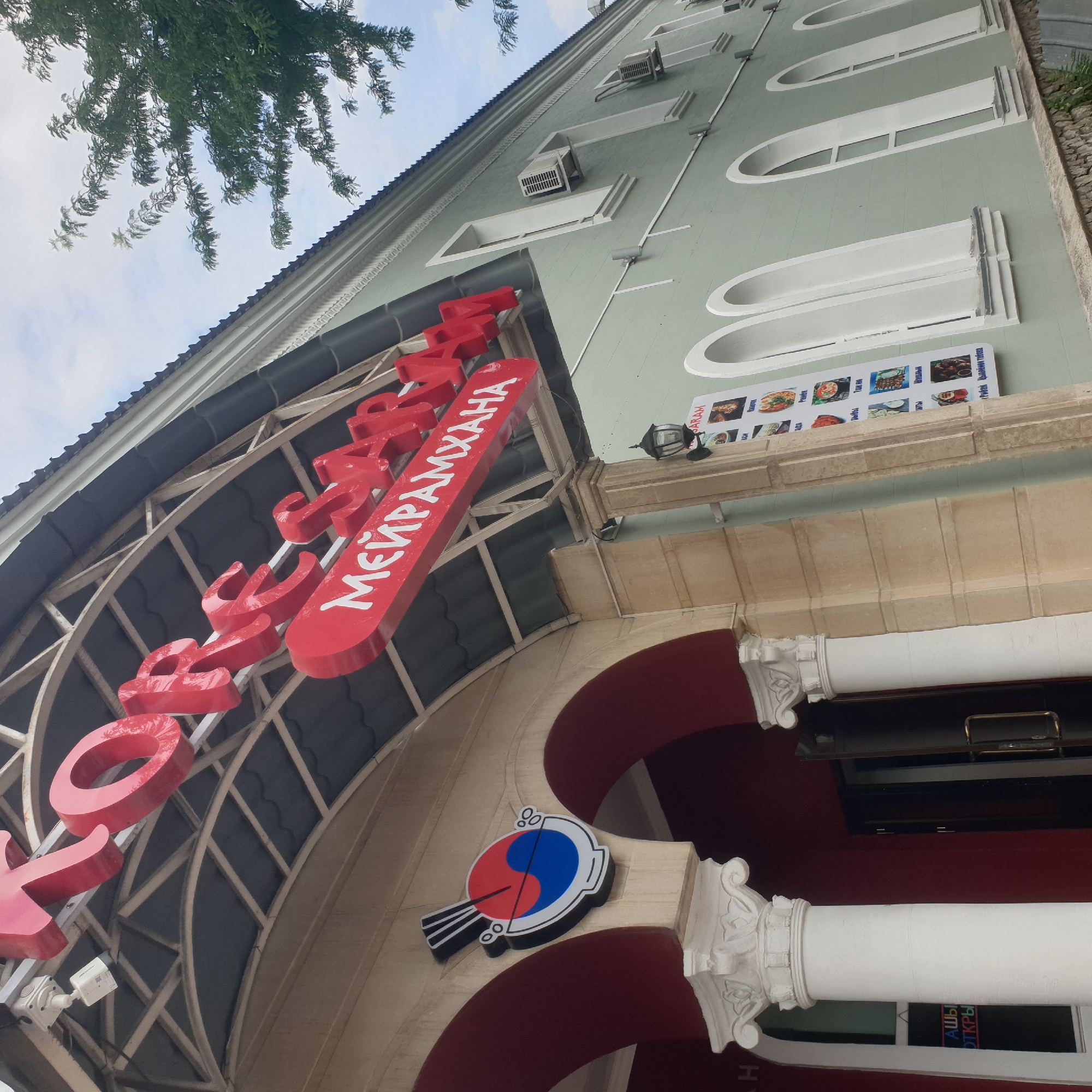 The image shows the exterior of a restaurant named "MEPAMXAHА" in Shymkent, Kazakhstan. The name is displayed in large, red, three-dimensional letters on the facade of a light green building. Above the entrance, a curved metal structure supports the signage. To the right of the entrance, there's a menu board with pictures of various dishes and their names in what appears to be Cyrillic script, alongside a logo resembling the Korean yin-yang symbol.
The building has a classical architectural style, with decorative white columns and arched windows. The upper part of the building shows air conditioning units installed on the wall. The sky is visible, suggesting it's daytime, possibly overcast due to the diffused light. A tree with green foliage partially obscures the top left of the image. Security cameras are also visible on the building's facade, indicating a concern for safety.
The overall scene depicts a commercial establishment, likely a restaurant, with clear signage and promotional materials aimed at attracting customers. The presence of specific dishes on the menu might hint at the type of cuisine offered, possibly influenced by Asian flavors given the yin-yang symbol. The image captures a street-level view of the building, focusing on the entrance and prominent signage.