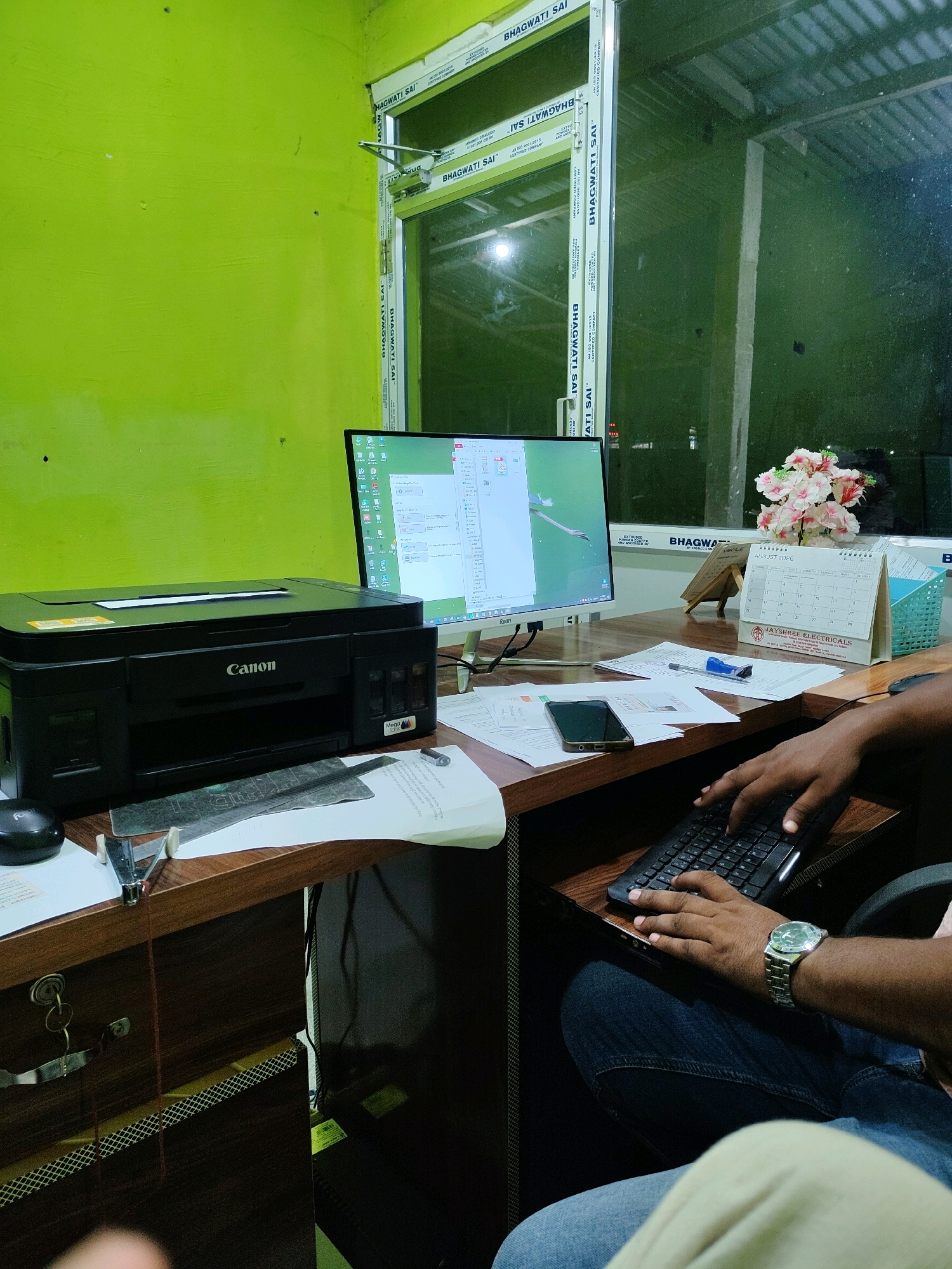 The image shows an indoor office scene. A person's hands are visible typing on a black keyboard, wearing a silver watch on their left wrist. The desk is made of wood and contains a black Canon printer, a computer monitor displaying a document, scattered papers, a stapler, a mobile phone, and a calendar showing "AUGUST 2026". A bouquet of pink and white flowers sits to the right of the monitor. The wall behind the desk is painted bright green, and a large window with "BHAGWATI SAI" lettering on the frame overlooks an outdoor area with a corrugated metal roof. The scene is illuminated by artificial lighting. There is no explicit information or visual cues indicating the location is specifically in Silbori, India.
