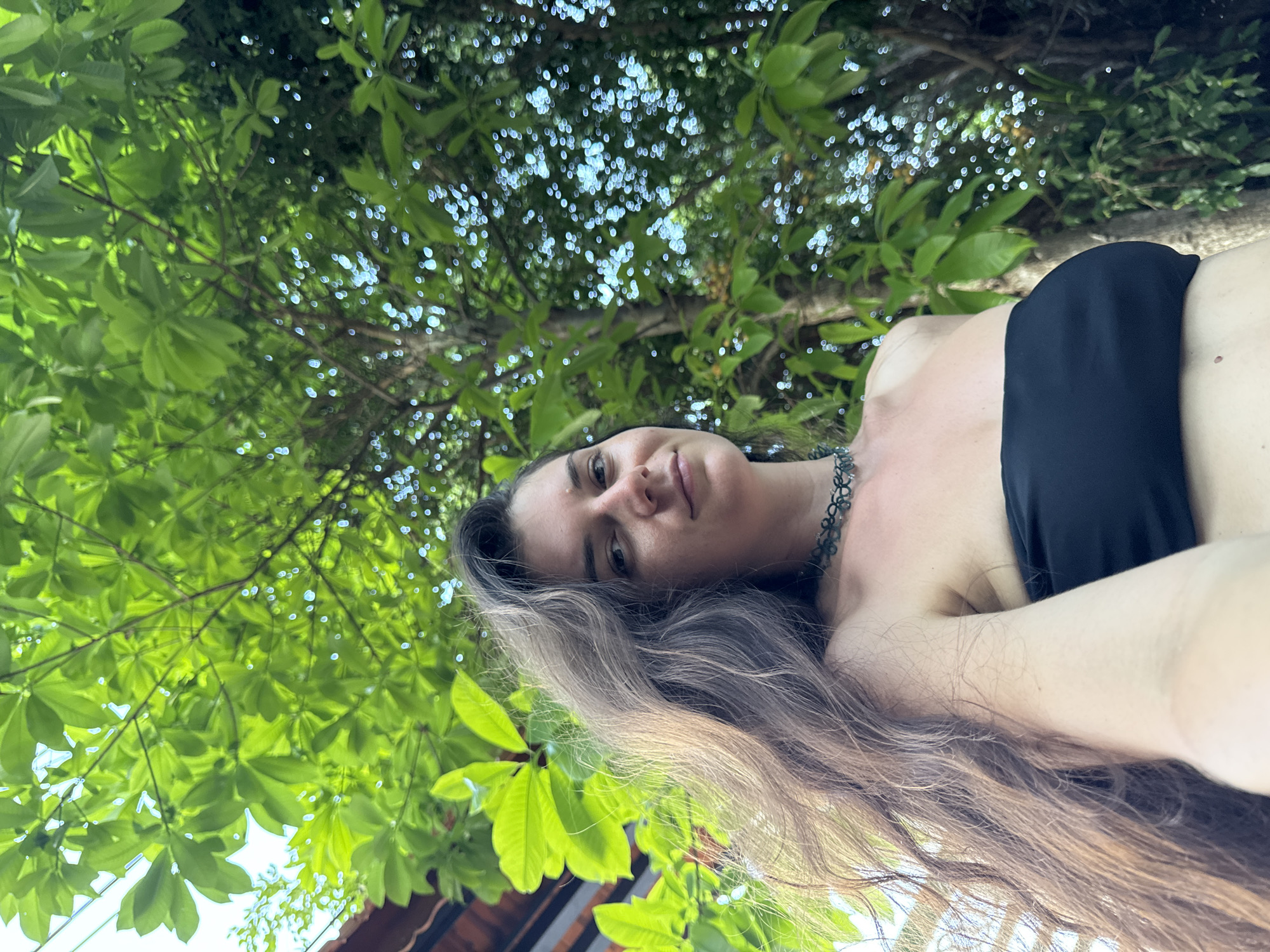 A low-angle, portrait-oriented photograph features an adult female outdoors in Mũi Né, Vietnam, set against a backdrop of lush green foliage and bright daylight. The subject, positioned centrally and looking directly at the camera, has long, wavy, medium-brown hair that falls over her left shoulder and chest. She is wearing a black strapless bandeau top and a dark green, choker-style necklace with a delicate, possibly leaf-like pattern. Her right arm is partially visible, extending towards the lower frame. The background is dominated by dense green leaves and tree branches, with sunlight filtering through, creating luminous bokeh effects and bright highlights, particularly on the right side. In the upper left corner, a portion of a building with a reddish-brown tiled roof and wooden eaves is visible, partially obscured by the foreground foliage.