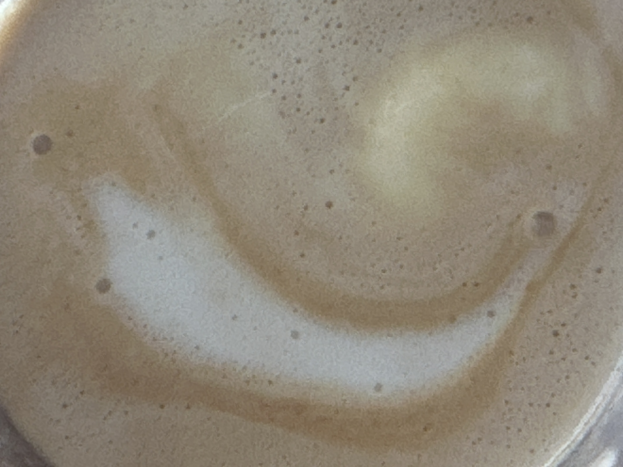 The image displays a close-up, overhead perspective of the surface of a frothy, light brown beverage, consistent with a coffee drink such as a latte or cappuccino. The surface is textured with numerous small bubbles, indicative of frothed milk or crema. A distinct, lighter, off-white, crescent-shaped swirl is visible in the central-left area, suggesting simple latte art or the natural mixing of ingredients. Scattered across the brown froth are several darker, small circular depressions. The color palette is dominated by shades of beige, light brown, and off-white. A minimal portion of the container's rim is discernible along the bottom edge of the frame. This beverage is presented in the context of being located in Fethiye, Turkey. No people, discernible actions, or environmental details beyond the drink itself are present in the image.