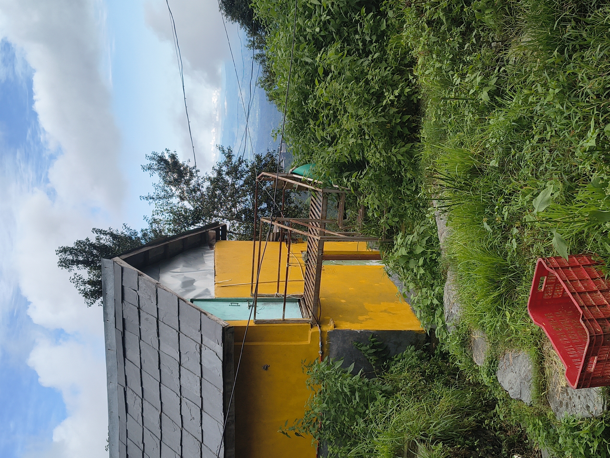 The image depicts a yellow building with a dark grey, slate-tiled roof. A rusty metal staircase leads to a small balcony or platform adjacent to a turquoise door. The building is situated on a hillside with dense green vegetation surrounding it. In the foreground, a red plastic crate rests on a stone path amidst the grass. In the background, a valley with distant mountains and scattered clouds can be seen under a blue sky. The scene is outdoors, likely in a rural or mountainous area, with elements suggesting a dwelling or outbuilding. There are no people or animals visible in the image. The location is identified as Haini, India.