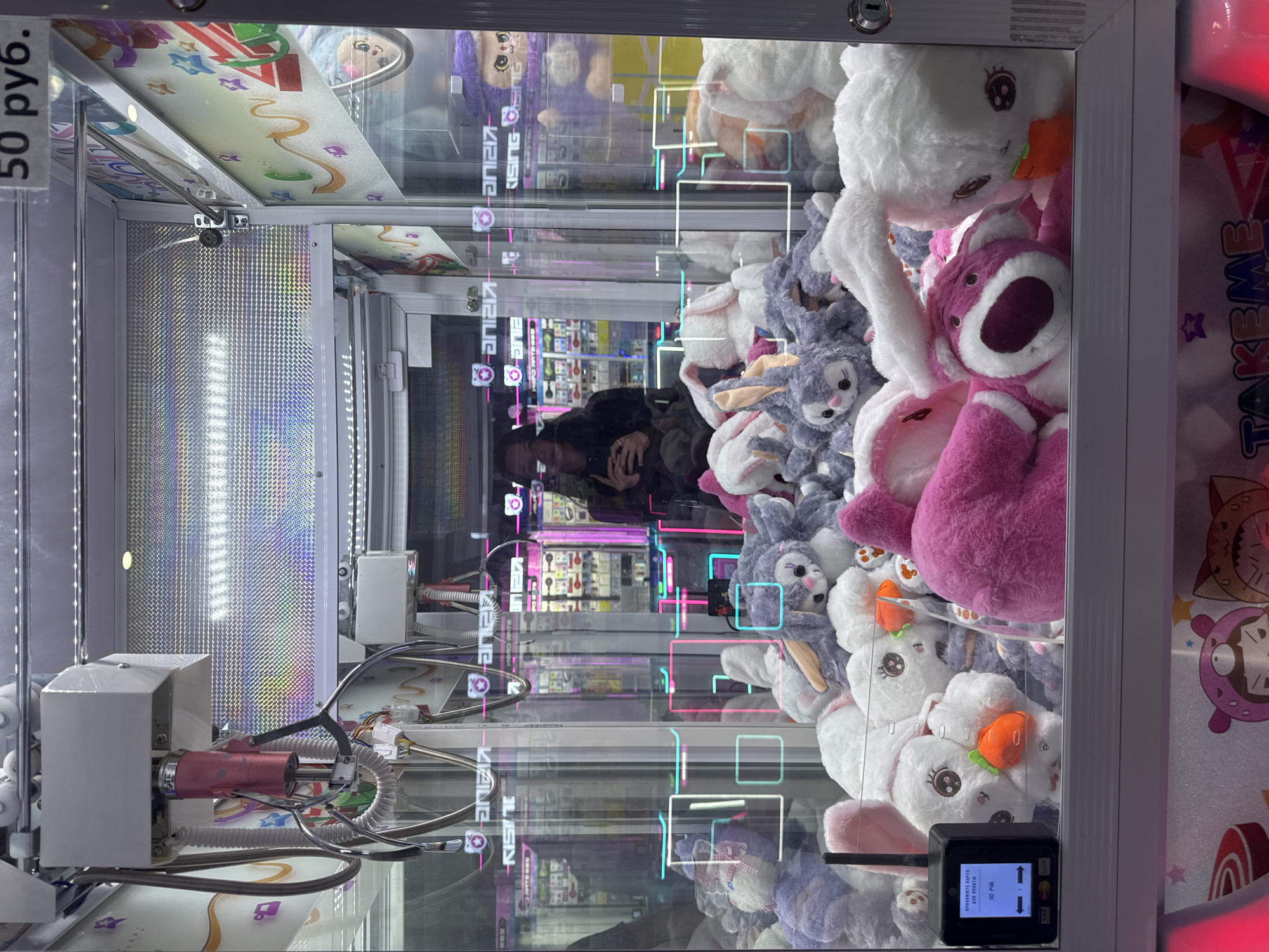 The image is a medium shot of a claw machine filled with plush toys, primarily rabbits and a pink bear character. The machine is located in Sochi, Russia, as indicated by the visible Cyrillic text "50 руб." (50 rubles) at the top right of the image, which is a common currency in Russia. The claw mechanism is visible at the top of the machine, along with a display screen on a small black device on the left, showing "50 РУБ." and other text. A person's reflection is visible in the glass, holding a phone, suggesting they are taking the photo or interacting with the machine. The background shows the interior of what appears to be an arcade or entertainment center, with other similar machines visible, also illuminated with colorful lights. The overall scene depicts a typical setup for a claw machine prize game.
