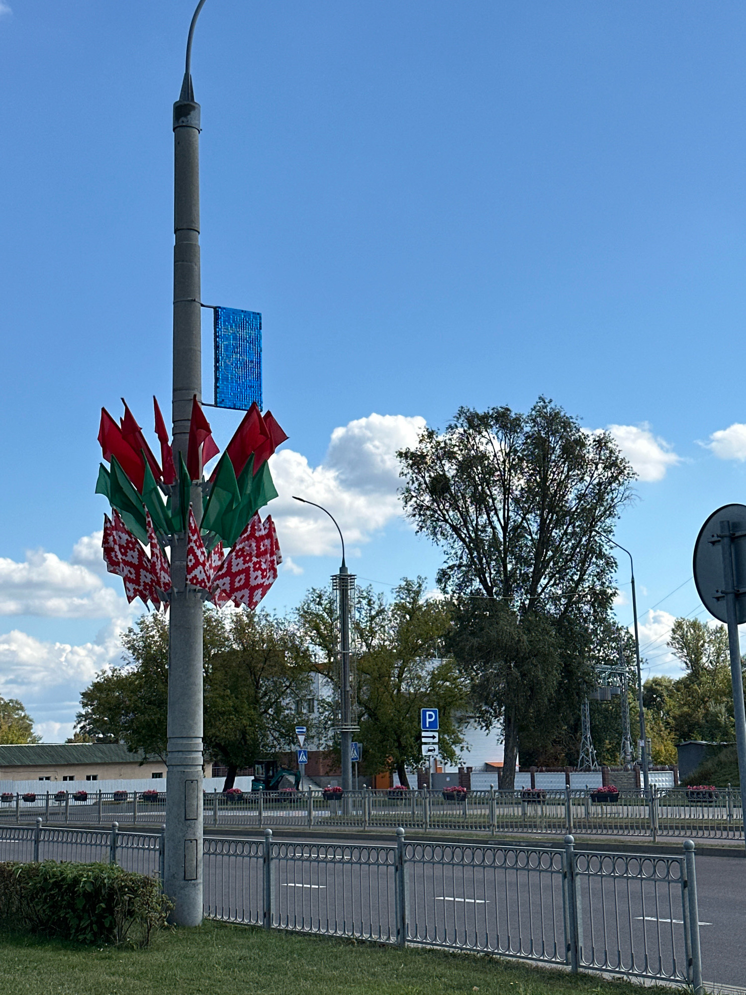 The image depicts a street scene in Brest, Belarus, under a bright blue sky with scattered clouds. A prominent lamppost on the left side of the frame is adorned with numerous small flags. These flags are arranged in a circular pattern around the lamppost and feature a repeating geometric pattern in red and white, alternating with solid red and green flags. Above the flags, a blue, rectangular object, possibly a solar panel or a decorative light fixture, is attached to the lamppost.
In the background, a paved road runs horizontally across the image, separated from the sidewalk by a decorative metal fence. Beyond the road and fence, several buildings are visible, including a long, low structure with a green roof on the left and other buildings further in the distance. Trees with green foliage are scattered throughout the background, with a particularly large, leafy tree dominating the right side of the frame. Traffic lights and a parking sign are also visible in the distance. The overall impression is a public space decorated for an event or celebration.