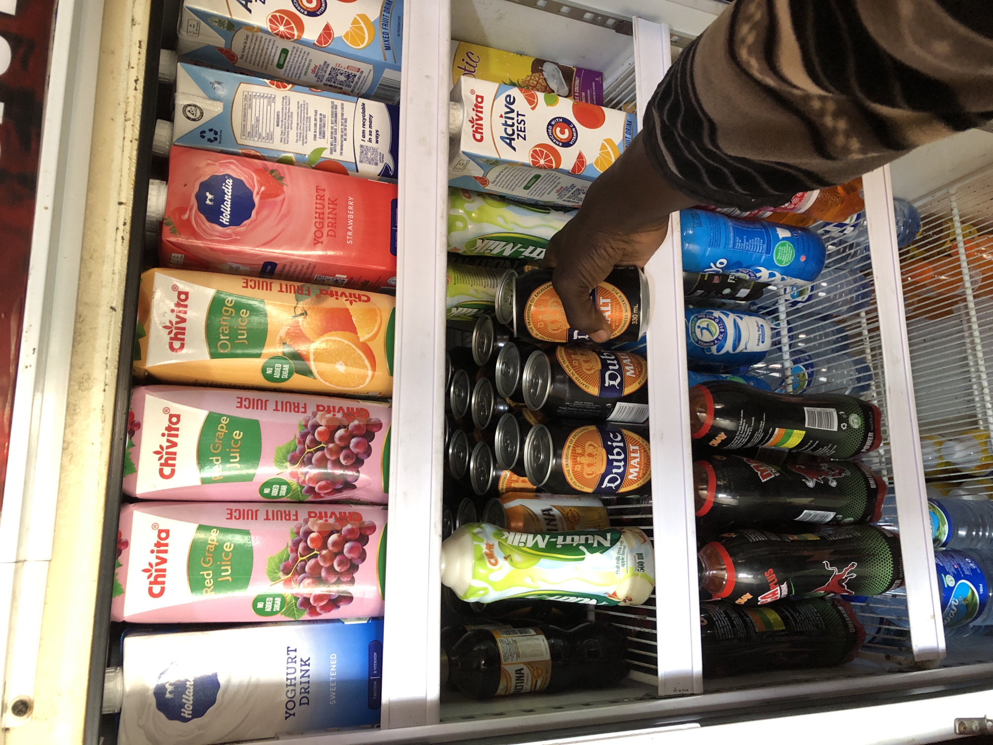 The image shows the inside of a commercial refrigerator stocked with various beverages. The refrigerator is located in a shop in Jos, Nigeria. The top shelf displays several cartons of juice, including "Chivita Red Grape Juice" and "Chivita Orange Juice," as well as a "Hollandia Yoghurt Drink." Below this, the middle shelf holds a variety of canned drinks, such as "Dubic Malt" and "Nutri-Milk." A person's hand is reaching into the refrigerator, holding a can of "Dubic Malt." The bottom shelf is filled with bottles, including energy drinks and water. The overall environment appears to be a retail setting, likely a convenience store or a similar establishment.