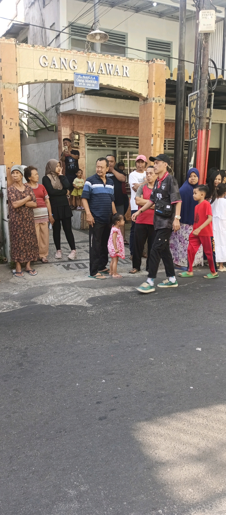 A group of people are gathered near the entrance to "Gang Mawar" in Malang, Indonesia. The entrance is marked by an arched gateway with "GANG MAWAR" written in raised lettering. A blue street sign below reads "JL. NAKULA GANG. MAWAR RT. 01 - RW. 05". On the right side of the gateway, another sign vertically displays "GANG MAWAR". A streetlamp hangs from the top of the gateway.
Several individuals are standing on the sidewalk and the street. On the left, three women are standing together, with one in the foreground wearing a patterned dress and sandals. Next to her, a woman in a red top and patterned pants stands with her arms crossed. Behind them, a woman in a black hijab and pants looks to the side.
In the center of the image, a group of men and children are present. A man in a blue and white striped shirt is looking to the right. A young girl in a pink floral dress stands near his feet. To his right, a man in a black t-shirt and cap is walking away from the camera. Behind him, a man in a red vest and black pants stands with his arms crossed. A young boy in a yellow shirt is visible further back.
On the right side, several women are gathered. One woman wears a blue hijab and a patterned skirt. A young boy in a red shirt and pants stands to her right, looking to the left. Another woman in a white t-shirt and pink cap is also in this group.
The environment appears to be a street with asphalt pavement. The buildings visible have brick or tiled exteriors. Electrical wires are strung across the scene, and a utility box is mounted on a pole. The lighting suggests it is daytime, with some shadows cast on the ground.