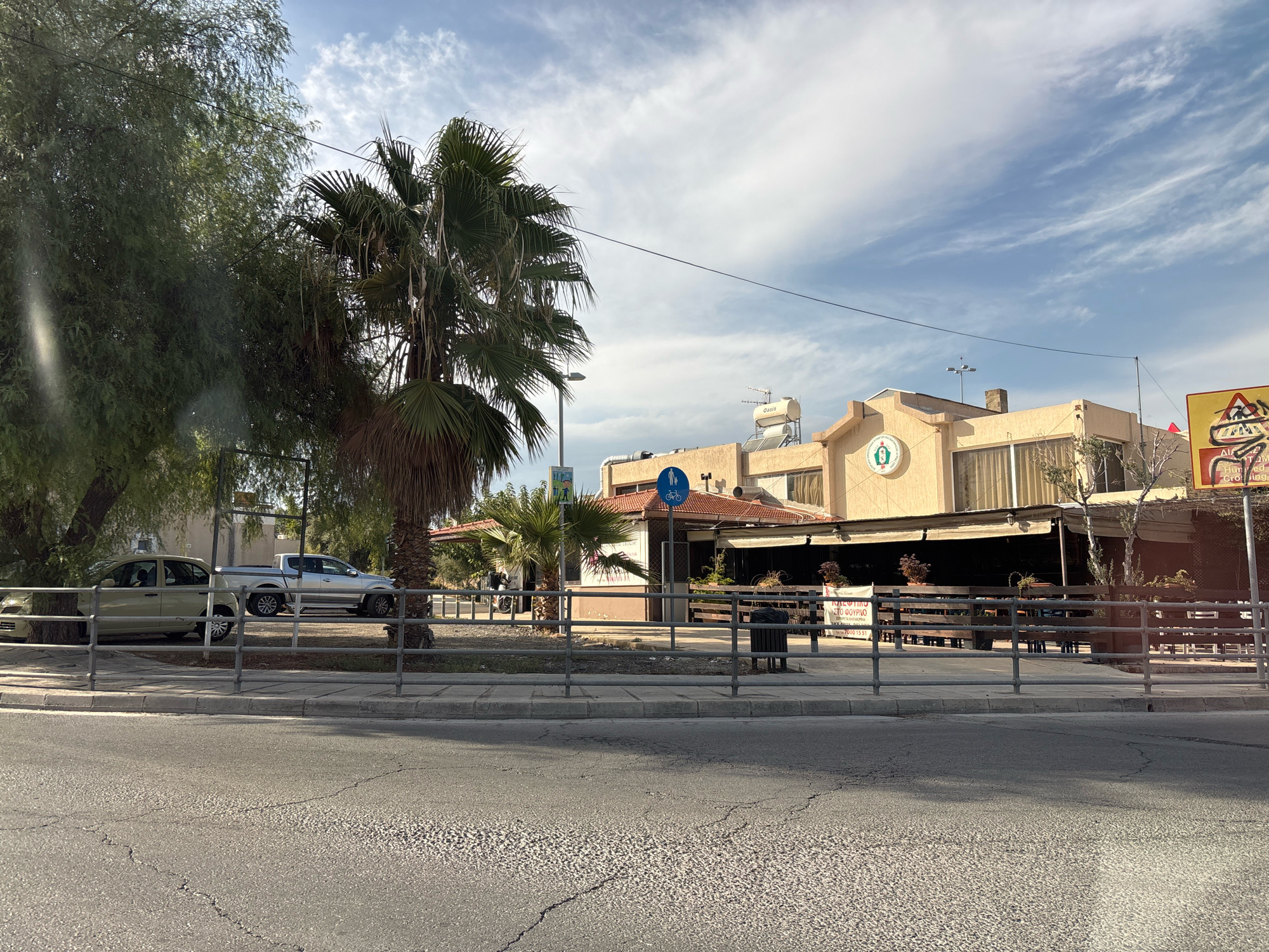 The image shows a street scene in Mesa Geitonia, Cyprus. In the foreground, a cracked asphalt road is visible. Behind the road, a grey metal fence separates the sidewalk from a paved area with a sidewalk. To the left, a light-colored car and a silver pickup truck are parked. Beyond the parked vehicles, a large tree with sparse foliage is present, and a palm tree stands to its right. In the background, a light-colored, two-story building with a tiled roof houses a business, possibly a restaurant or cafe, with outdoor seating visible. A blue sign with a pedestrian and bicycle symbol is mounted on a pole in front of the building. The sky above is partly cloudy with blue patches. Another building with a solar water heater is visible on top of the main building. A yellow triangular traffic sign with a red border and black symbols is positioned on the far right side of the image, depicting a pedestrian crossing.