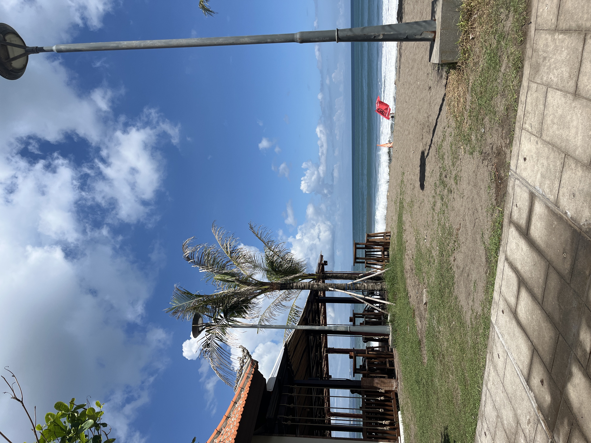 This is a landscape photograph taken in North Kuta, Indonesia. The image captures a coastal scene with a beach, ocean, and sky. In the distance, there's a red warning flag planted in the sand near the shoreline, along with an individual standing beside it. The beach transitions into a grassy area bordered by a stone walkway or pavement in the foreground.
A structure with a red-tiled roof and open-air seating is visible. A palm tree grows nearby, adding to the tropical atmosphere. Above, the sky is bright blue with scattered clouds, and a light pole cuts across the upper left portion of the frame.