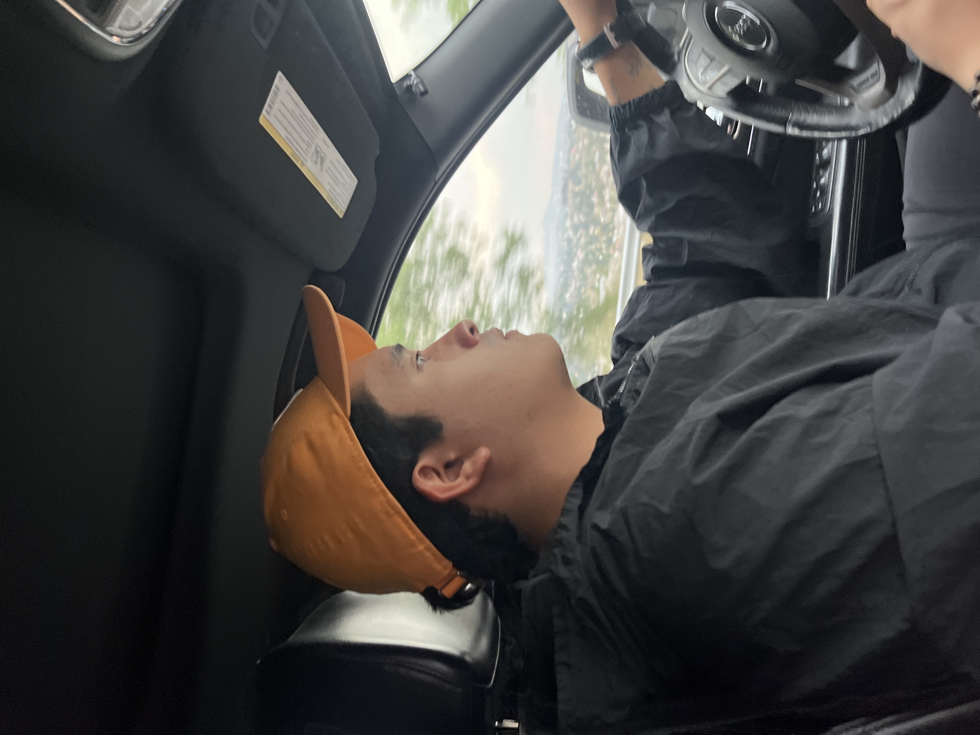 A man wearing an orange cap and a black jacket is seated in the driver's seat of a car. He is looking upwards and towards the right, in the direction of the car's windshield. His hands are on the steering wheel. The scene is inside the car, and outside the window, there is a blurry view of trees. The location is not discernible as Santa Clara Ocoyucan, Mexico based on the visual information provided.