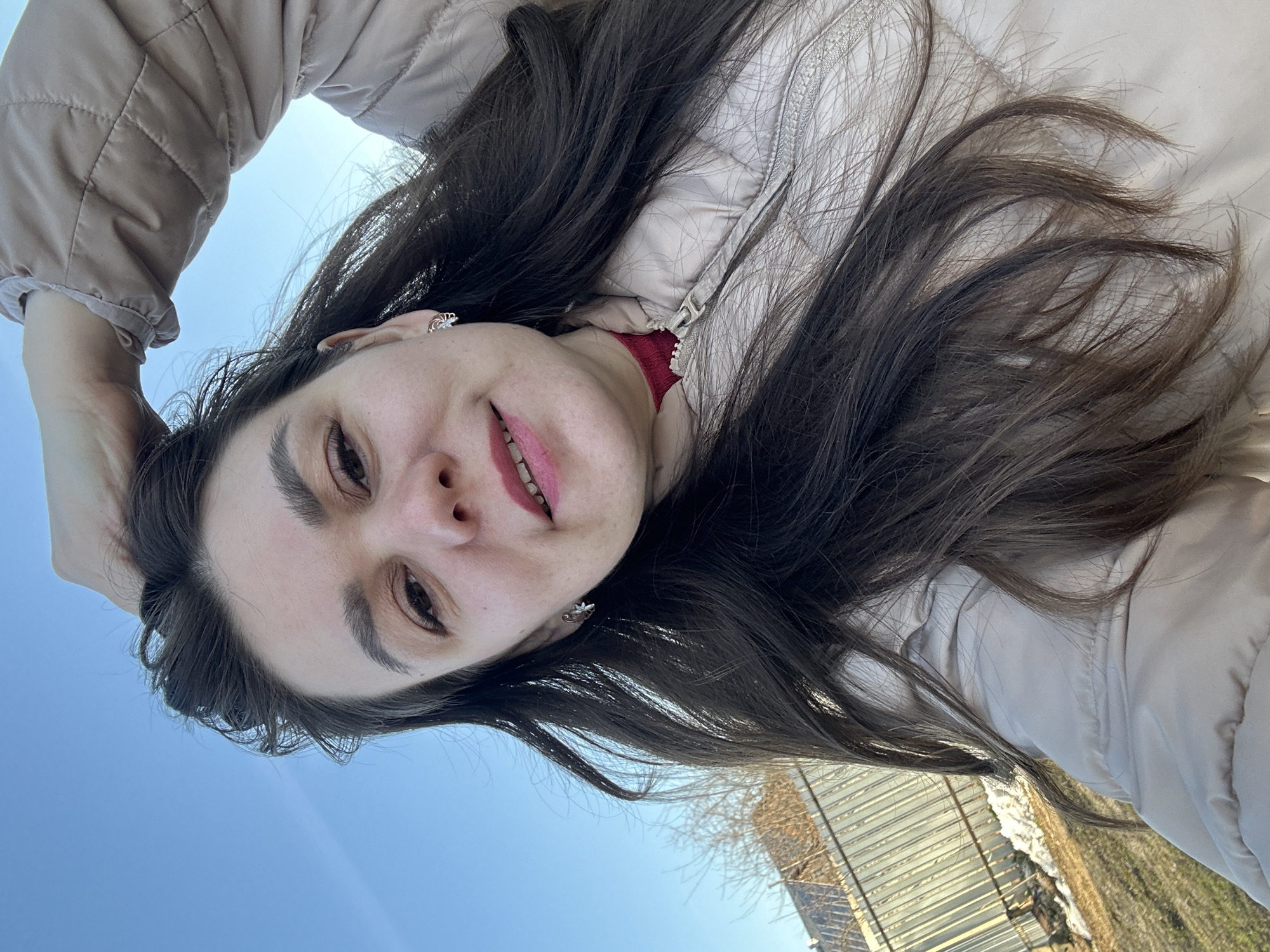 The image features a young woman in a low-angle, upward-facing shot, likely a selfie, taken outdoors in Chaikovsky, Russia. Her face, framed by long, dark brown hair, is the primary subject. She has dark eyebrows, brown eyes, and fair skin, and wears light-pinkish-red lipstick, revealing a gentle smile. She is adorned with small, silver-toned stud earrings, one resembling a crown-like design. Her attire includes a light beige or cream-colored quilted jacket, with a hint of a red garment visible underneath at her neck.
The background is dominated by a clear, bright blue sky, indicating a sunny outdoor setting during daytime. In the lower right corner, there are subtle hints of bare tree branches and a structure, possibly a fence or a building with a light-colored, possibly corrugated metal, roof or siding. The overall mood conveyed by her expression is cheerful and relaxed. No visible text is present in the image.