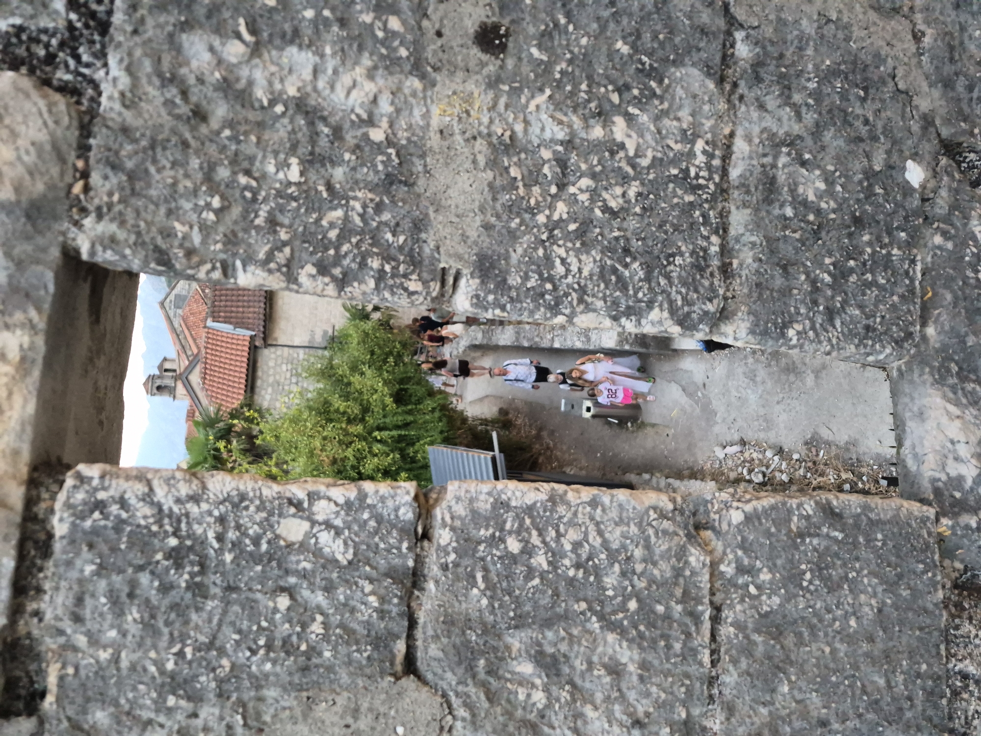 An outdoor, eye-level, medium shot captures a narrow walkway seen through a rectangular aperture in a rough stone wall in the city of Kotor, Montenegro. The walkway, lined with greenery on the left and more stone structures on the right, is populated by several individuals walking in the same direction. In the foreground of the walkway, a young girl in a pink t-shirt with the number "82" and white shorts is walking, followed by a woman in a white dress. Further up the walkway, a man in a white shirt and dark shorts is visible. Other individuals are further back in the frame, some appearing to be tourists. The architectural style of the buildings in the background, with their red-tiled roofs and stone construction, is characteristic of Kotor's old town. The lighting suggests it is late afternoon or early evening.
