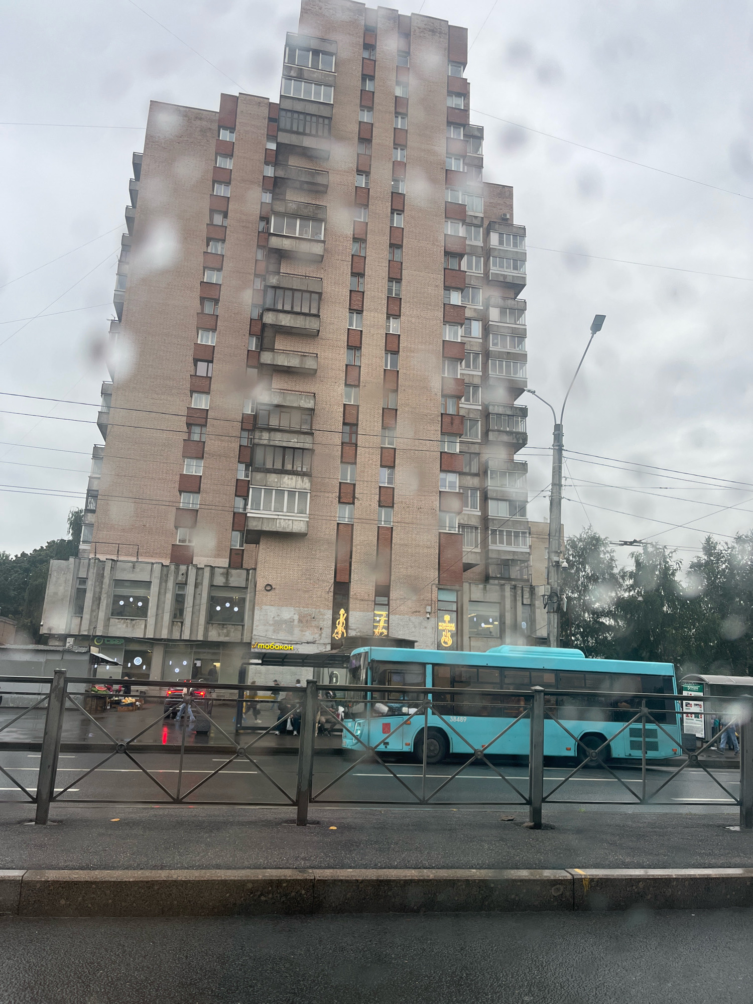 This image captures a street scene in Saint Petersburg, Russia, likely on a cloudy or rainy day, evidenced by water droplets on the camera lens. A tall, multi-story brick building with numerous windows and balconies dominates the background. In the foreground, a turquoise bus with the number "38489" is visible, driving along the street. A metal railing separates the sidewalk from the road. Several people are seen on the sidewalk near the bus stop. Businesses with signage are present at the base of the building. Trees and a street lamp are also part of the urban landscape. The overall impression is of a typical city street on an overcast day.