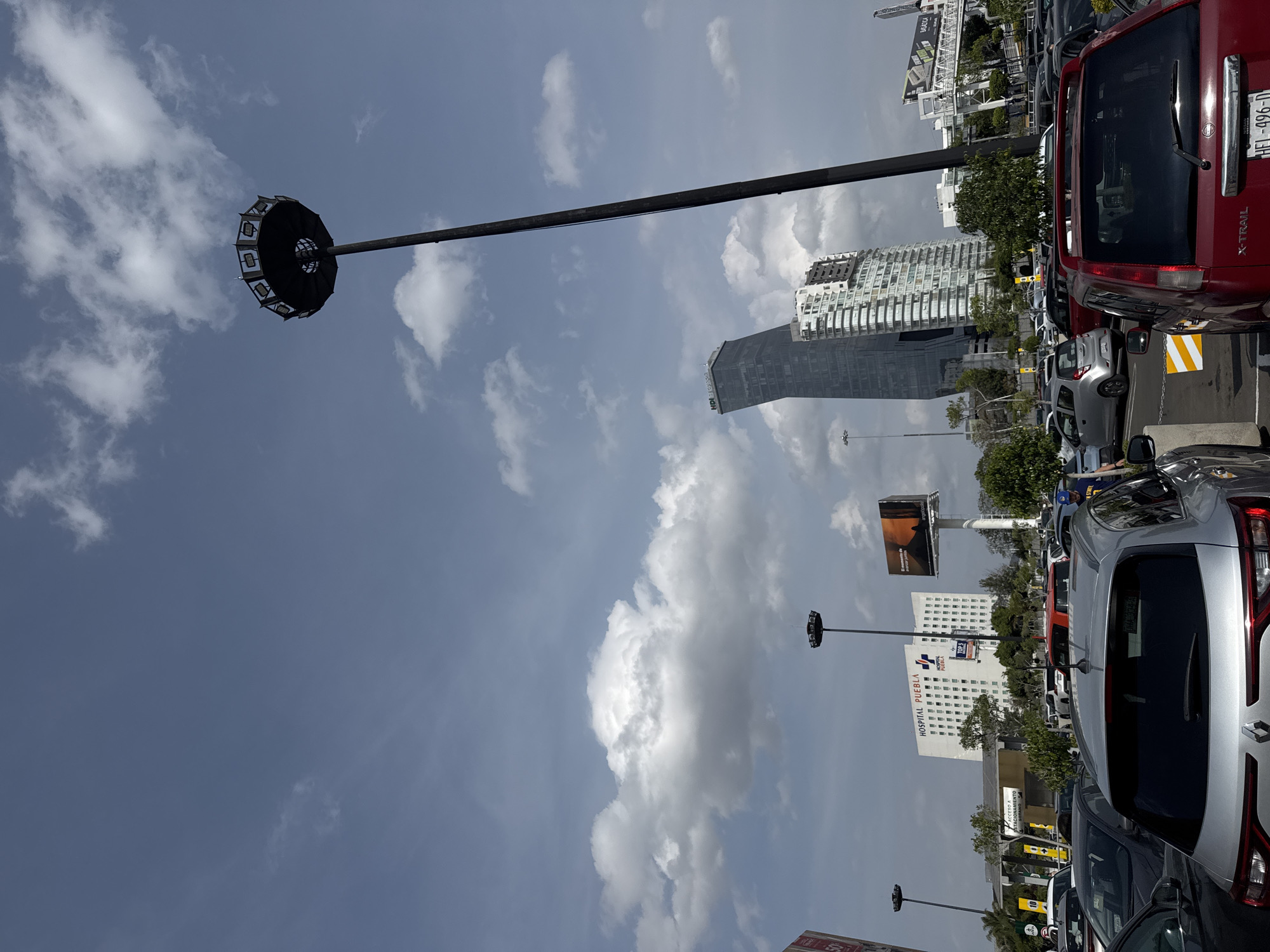The image shows a parking lot in Puebla, Mexico, with various vehicles, buildings, and a prominent light fixture. In the foreground, multiple parked cars are visible, including a red Nissan X-Trail with the license plate HEL-496-D. In the background, there is a building with "HOSPITAL PUEBLA" and "HOSPITAL" signage. Also visible are billboards, modern skyscrapers, and standard streetlights. A tall street lamp with a unique multi-bulb lantern fixture is positioned on the left. The sky is partly cloudy.