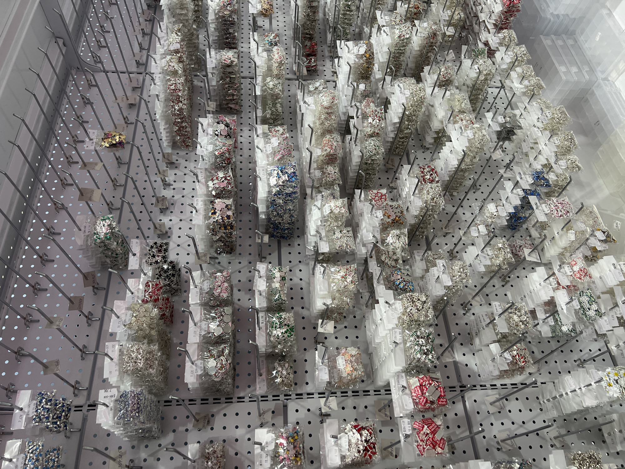 The image shows a display of various small decorative items, likely embellishments or craft supplies, hanging on a pegboard system. The items are packaged in clear plastic bags or small containers, and are arranged in rows and columns. The pegboard is metal and painted light gray or white. The items are of various colors and designs, with some appearing to be beads, sequins, or miniature fabric pieces. There are price tags or labels attached to some of the items. In the background, there are rows of clear plastic storage boxes. The scene appears to be in a retail or storage environment, such as a craft store. There is no evidence within the image to associate this with Burgas, Bulgaria.