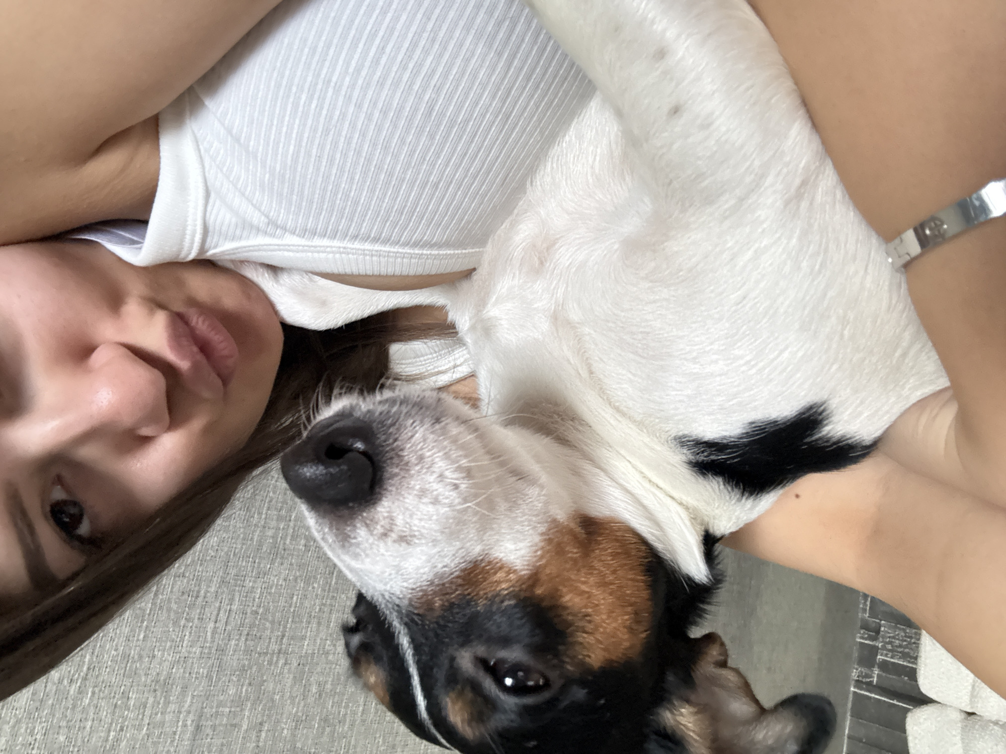 The image captures a close-up, rotated view of a person and a dog sharing a tender moment. A female individual, seen from the nose up to her upper chest, is positioned on the left side of the frame. She has light brown hair and is wearing a white, ribbed tank top. Her left arm is visible on the right, adorned with a silver bracelet that has faint, illegible markings. Her lips are slightly parted, and she looks towards the camera with a gentle, content expression.
Cuddled beside her on the right is a dog, likely a Jack Russell Terrier-type breed. The dog has prominent white fur with distinct brown and black patches around its eyes and ears. Its dark nose and one eye are clearly visible, conveying a calm demeanor. Both subjects appear to be resting on a light grey, textured surface, possibly a sofa or bed.
The lighting suggests an indoor setting, likely during the daytime, creating a soft and intimate atmosphere. The overall activity depicted is one of quiet companionship and affection between the owner and their pet. No external weather cues are present. The emotional tone is warm and peaceful. The context provided indicates the location is Sofia, Bulgaria.