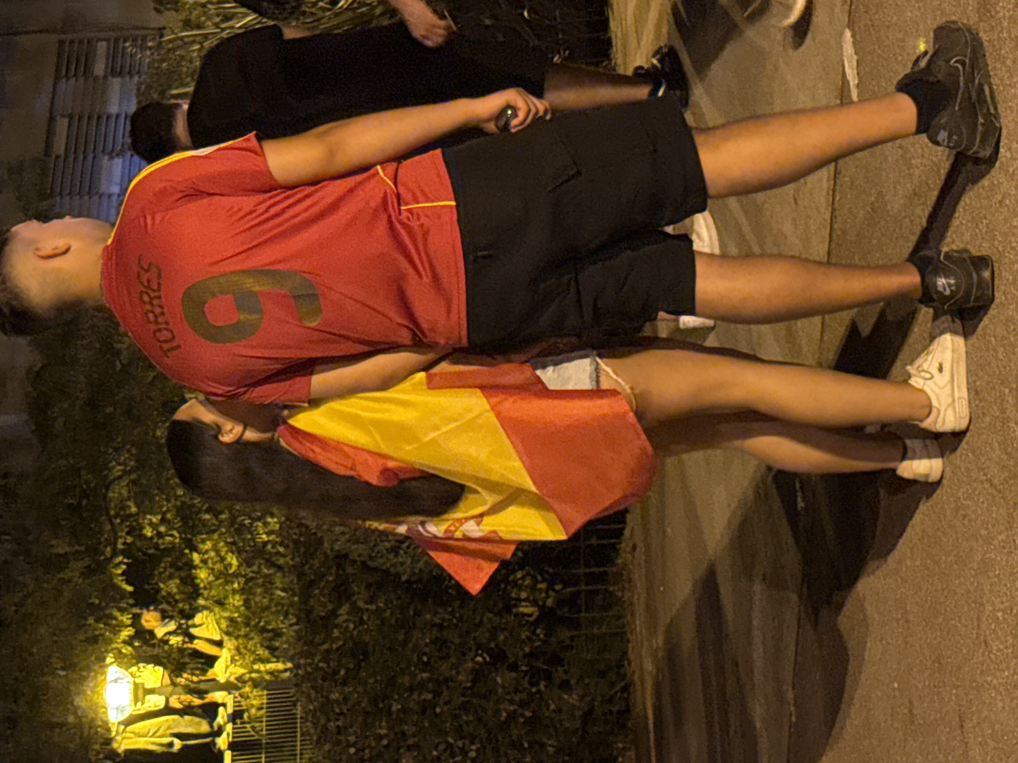 Two individuals, a woman and a man, are standing on a paved surface at night, with a Spanish flag draped over the woman's shoulders. The woman is on the left, facing away from the camera, and appears to be of Hispanic ethnicity with long, dark hair. She is wearing a light-colored garment and white sneakers. The man is to her right, also facing away from the camera. He is wearing a red sports jersey with the name "TORRES" and the number "9" on the back, black shorts, and black sneakers. A second man in a black t-shirt is partially visible behind and to the right of the man in the red jersey. In the background, there is dense foliage, a streetlamp with people standing near it, and a building with horizontal blinds. The scene appears to be outdoors, possibly on a street or promenade in Blanes, Spain, given the Spanish flag and the mention of the city. The lighting is dim, with the streetlamp providing the primary illumination.
