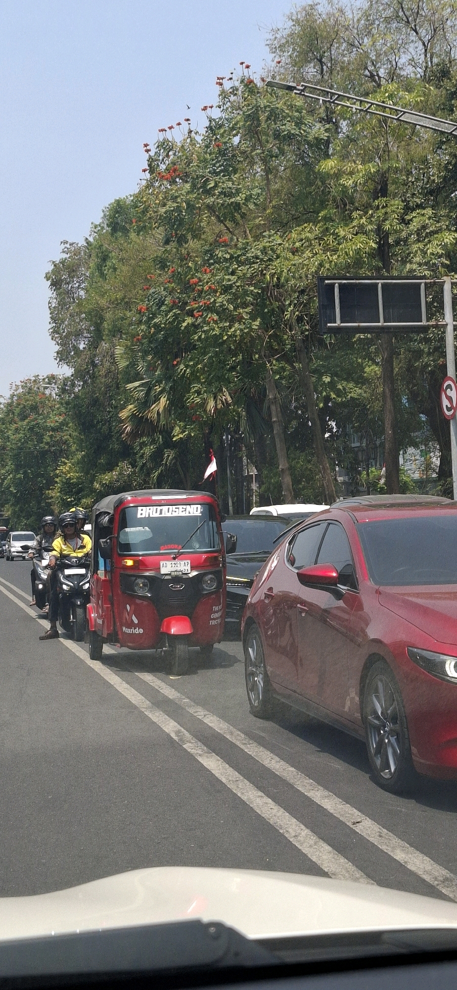 In a street scene in Surakarta, Indonesia, a red auto-rickshaw, identified as "BRUTUSEND" and bearing license plate "AD 1329 AP," is at the forefront, situated on the right side of the road. It is flanked by a red car on its right and a motorcycle carrying two individuals on its left. The auto-rickshaw displays the "Maxride" logo on its side. The road is marked with double white lines. Beyond the vehicles, a lush green canopy of trees with red flowers creates a vibrant backdrop, suggesting a location with significant vegetation within the city. Traffic lights and street signs are visible in the upper right quadrant of the image, indicating a typical urban traffic environment. The sky is clear and blue.