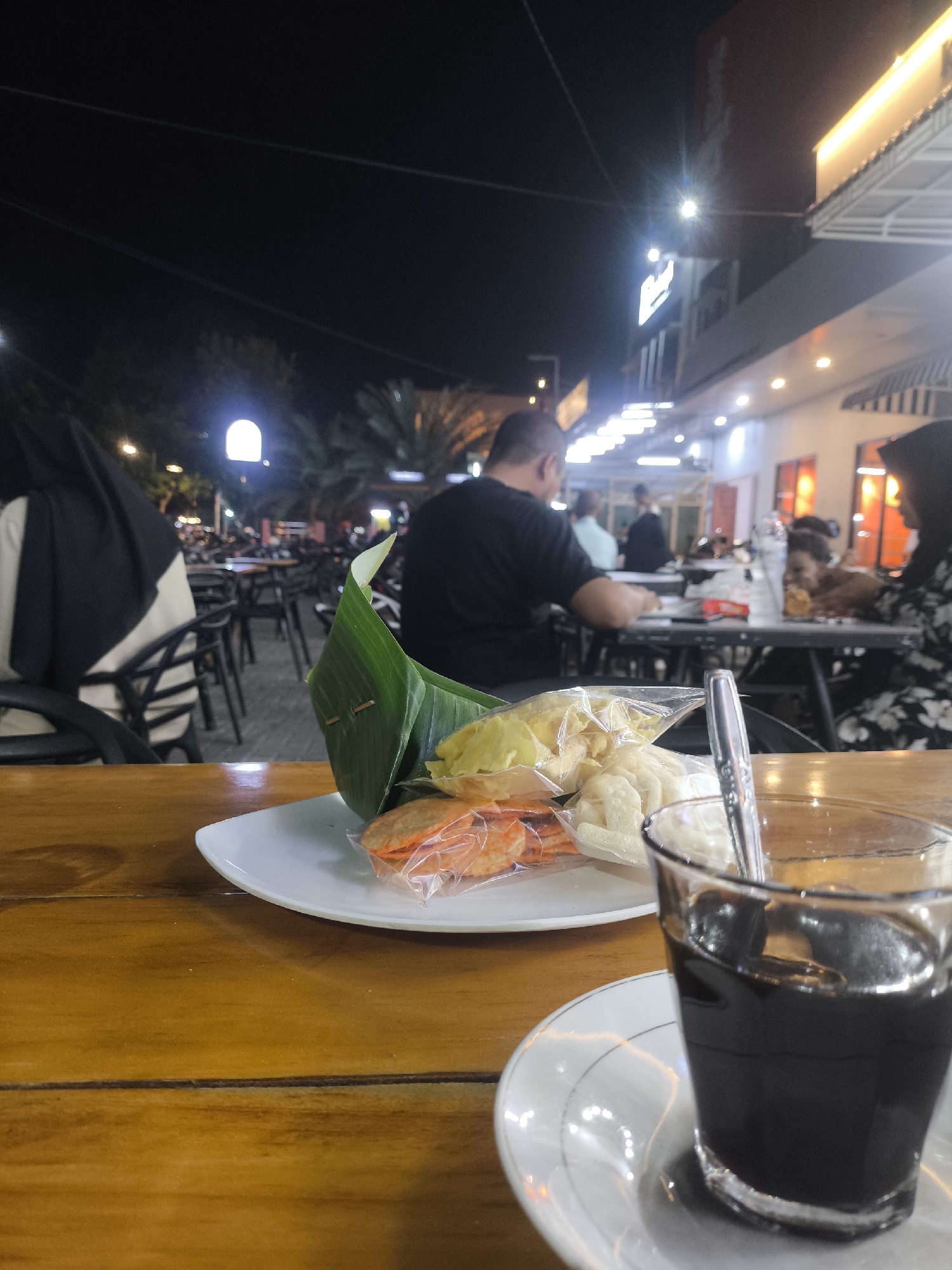 An outdoor dining scene at night in Bireuen, Indonesia, features a wooden table in the foreground with a plate of snacks and a glass of coffee. The snacks, wrapped in plastic and a banana leaf, include what appear to be crackers and fried items. A metal spoon rests in the coffee. In the background, people are seated at tables, with a blurred view of buildings and streetlights. One person in the foreground is wearing a black hijab. The overall atmosphere suggests a casual evening meal or gathering.