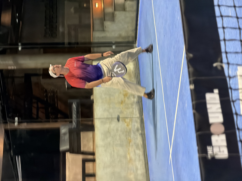 An adult male is observed on a blue padel court at "The Ground" facility in Pecatu, Indonesia, under artificial nighttime lighting. The Caucasian male, wearing a white baseball cap, eyeglasses, a red-to-purple gradient athletic shirt, khaki trousers, and dark athletic shoes, stands in an active athletic stance with bent knees and spread feet. He holds a dark padel racket, partially revealing "VAIRO" branding, in his right hand. His head is tilted back, and his gaze is directed upwards and to his left, indicating focus on an airborne object. A black net with a white "THE GROUND." logo band occupies the lower foreground. The background features a modern, enclosed or semi-enclosed structure composed of light-toned concrete walls, large glass panels, dark metallic support beams, and a visible staircase. Dark seating furniture is dimly visible through the glass panels, suggesting an adjacent viewing or lounge area.