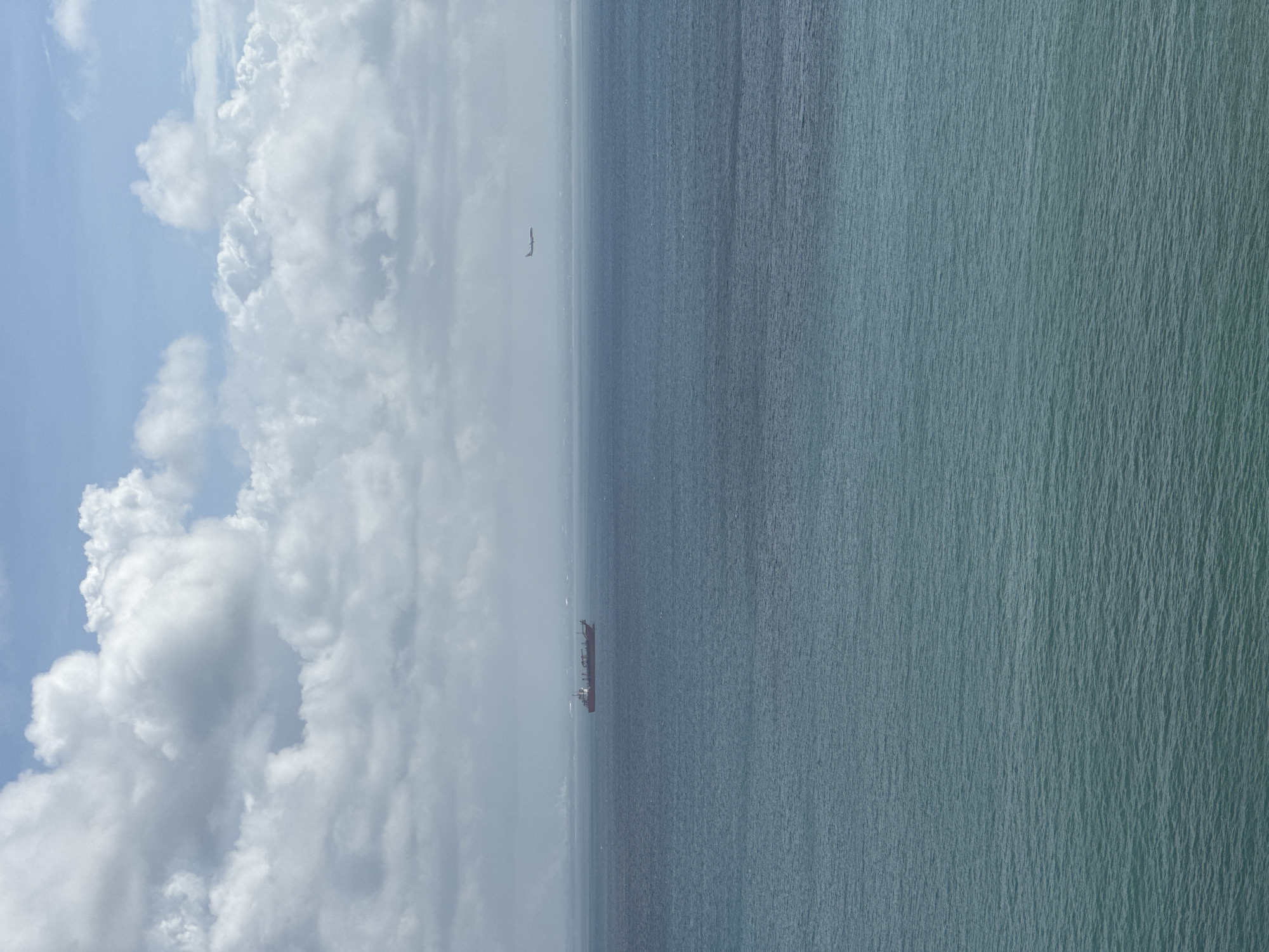 The image shows an aerial view of the ocean off the coast of Kuta Selatan, Indonesia. A cargo ship is visible in the center of the frame, moving horizontally across the image. A single bird is also visible in the sky. The sky on the left is filled with thick, white clouds under a blue sky. The ocean is a deep, aqua-green color.
