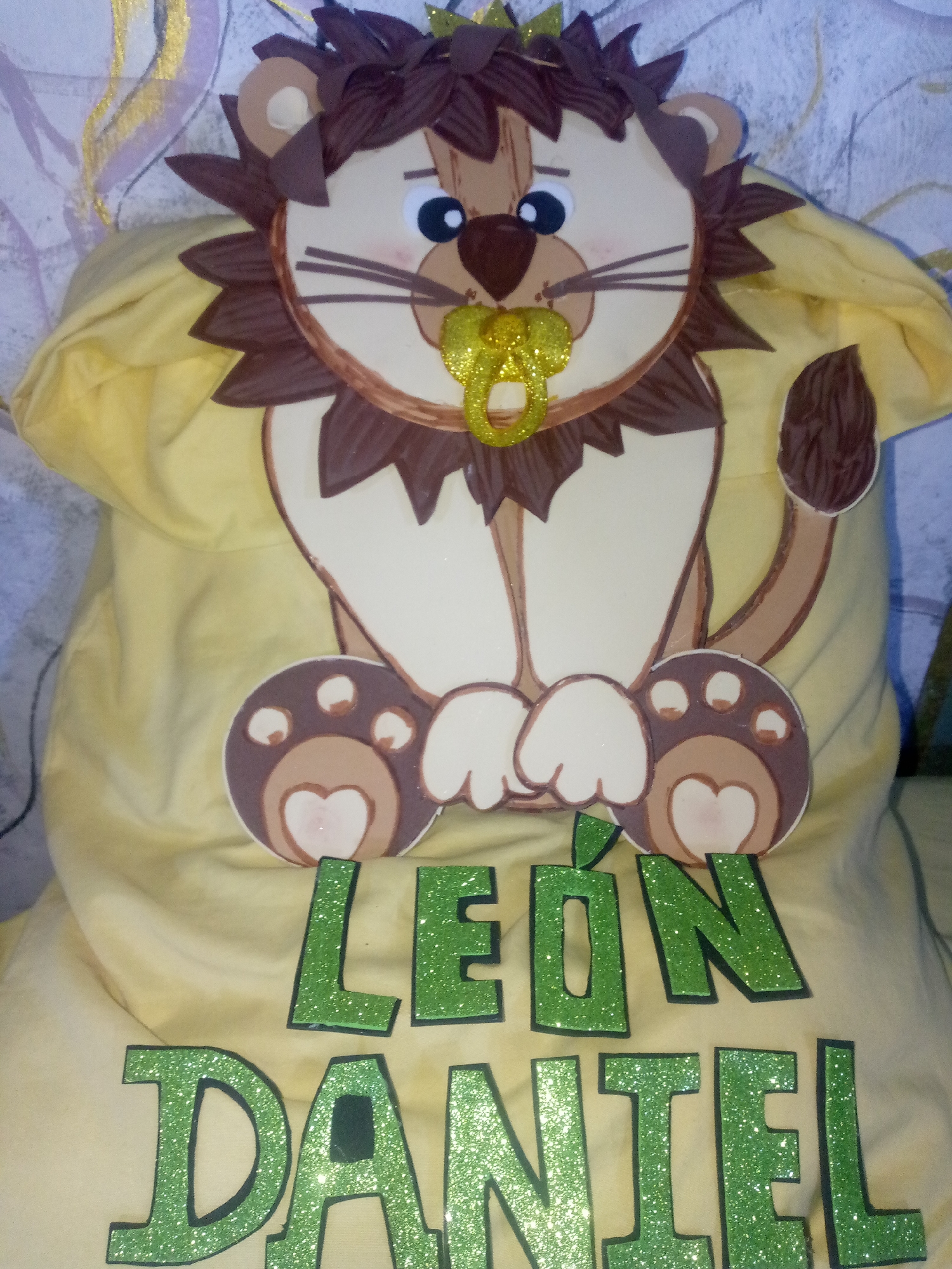 A yellow t-shirt featuring a cartoon lion and the name "León Daniel" in green glitter lettering is the primary focus. The lion illustration has brown fur, a tan face, a pacifier, and paw prints with heart-shaped pads. The lettering appears to be glued onto the shirt. The t-shirt is draped over an unseen surface, potentially a pillow or other soft item. The background consists of a mottled wall with a hint of gold paint. There is no information available to confirm the image location in the city of Juan Griego, Venezuela.