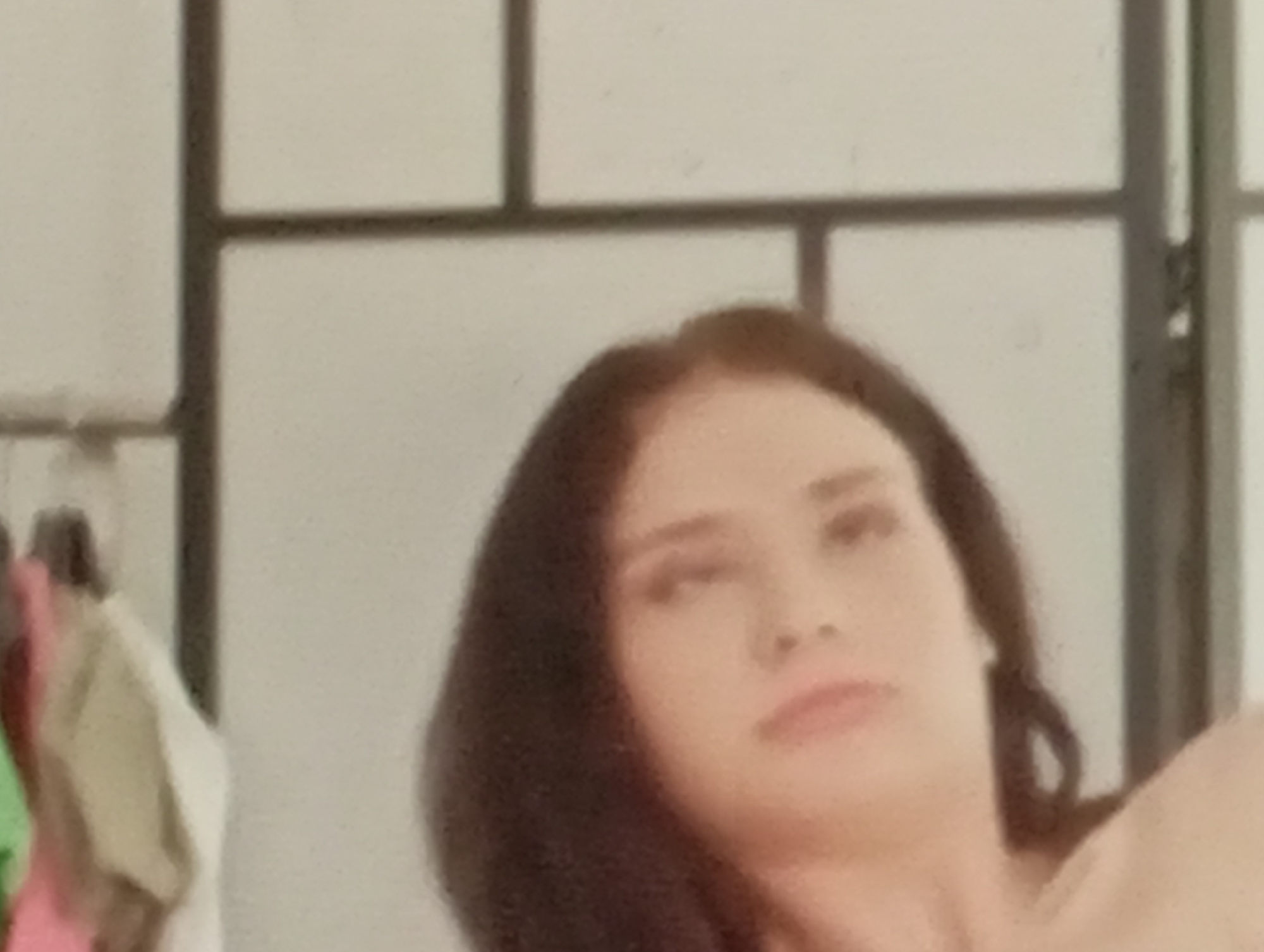 The image shows a close-up of a woman's face and upper body. She has long, dark hair, light-colored skin, and appears to be looking upwards and to the right. Her lips are slightly parted, and her eyes are open. The background is out of focus and appears to be a room with a light-colored wall and vertical dividers, possibly part of a screen or partition. There are some clothing items hanging on a rack to the left of the woman, but they are also out of focus. There is no discernible connection to Sofiivska Borshchahivka, Ukraine, in the image itself.