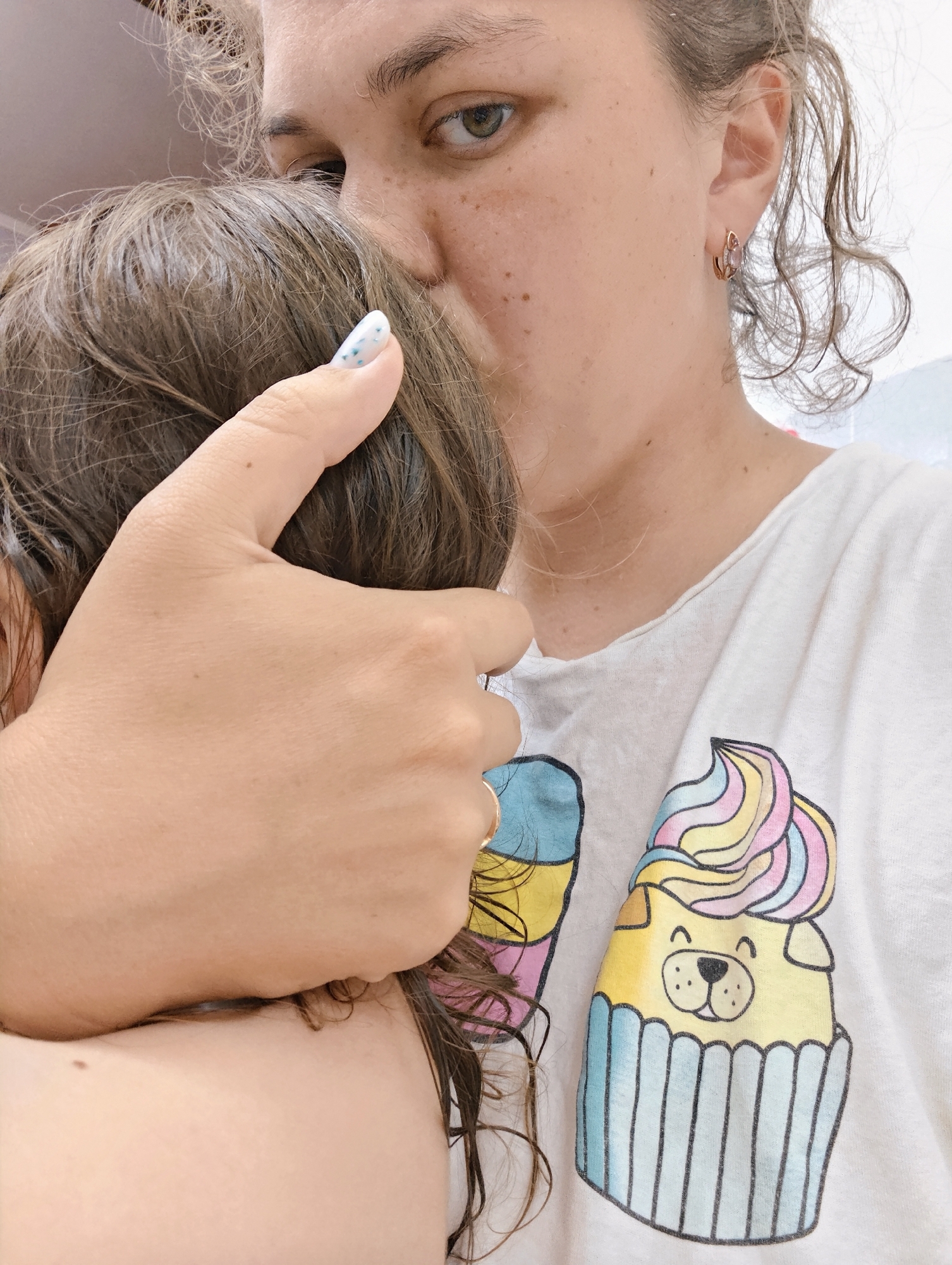 A person with light skin, freckles, and brown curly hair, wearing a white t-shirt with a cartoon cupcake design, is holding a child with brown hair. The person is looking towards the viewer. The scene likely takes place indoors, but the exact location within the city of Chaikovsky, Russia, cannot be determined from the image.