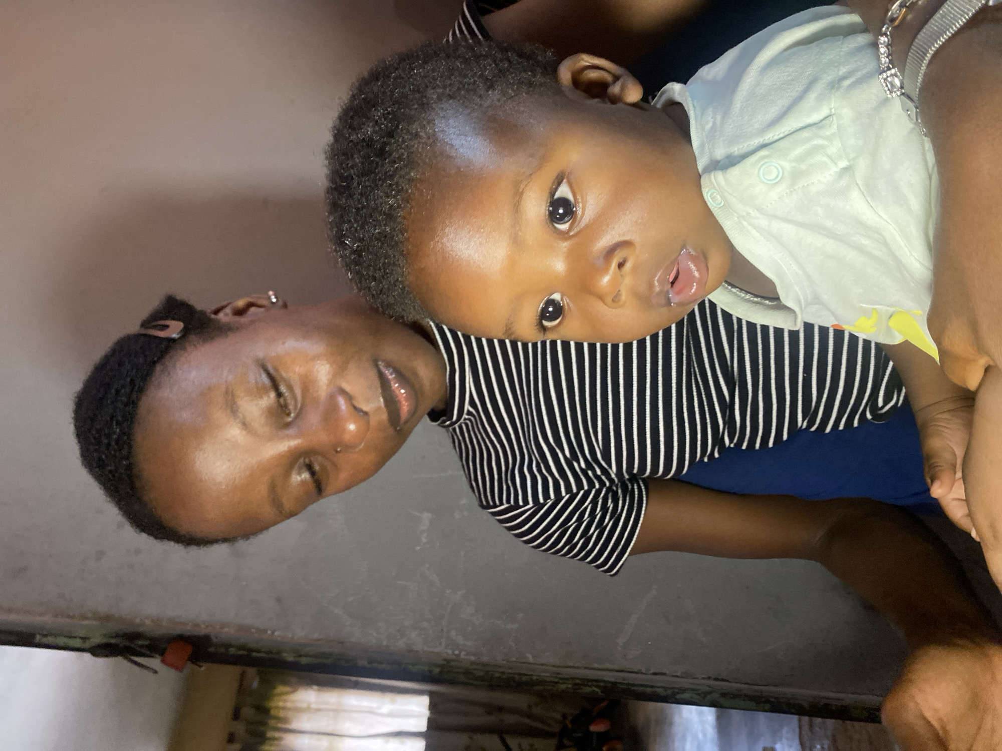 A close-up, eye-level shot captures a young Black woman holding a baby in Sabon Tasha, Nigeria. The woman, with braided hair pulled back, is looking down at the baby with a gentle expression. She is wearing a black and white striped, high-necked shirt. The baby, with wide, dark eyes and an open mouth, is looking forward, its head tilted slightly. The baby is dressed in a light blue, short-sleeved onesie adorned with a yellow graphic. The background is dimly lit, with a textured gray wall on the right and a window with sheer curtains visible on the left, suggesting an indoor setting. The overall scene conveys a moment of tender connection between the two individuals.