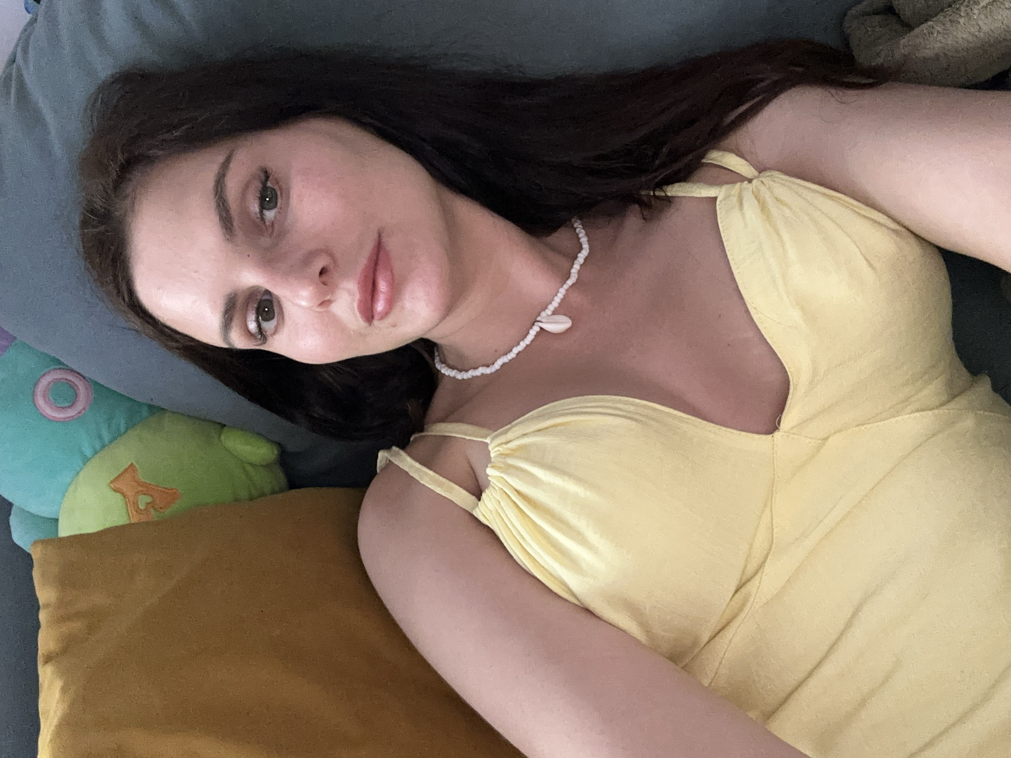 A woman with long, dark hair lies on a couch with a blue cushion. She is wearing a yellow dress and a shell necklace. To her left, a large orange pillow and a green stuffed animal are visible. The woman is looking at the camera with a neutral expression.