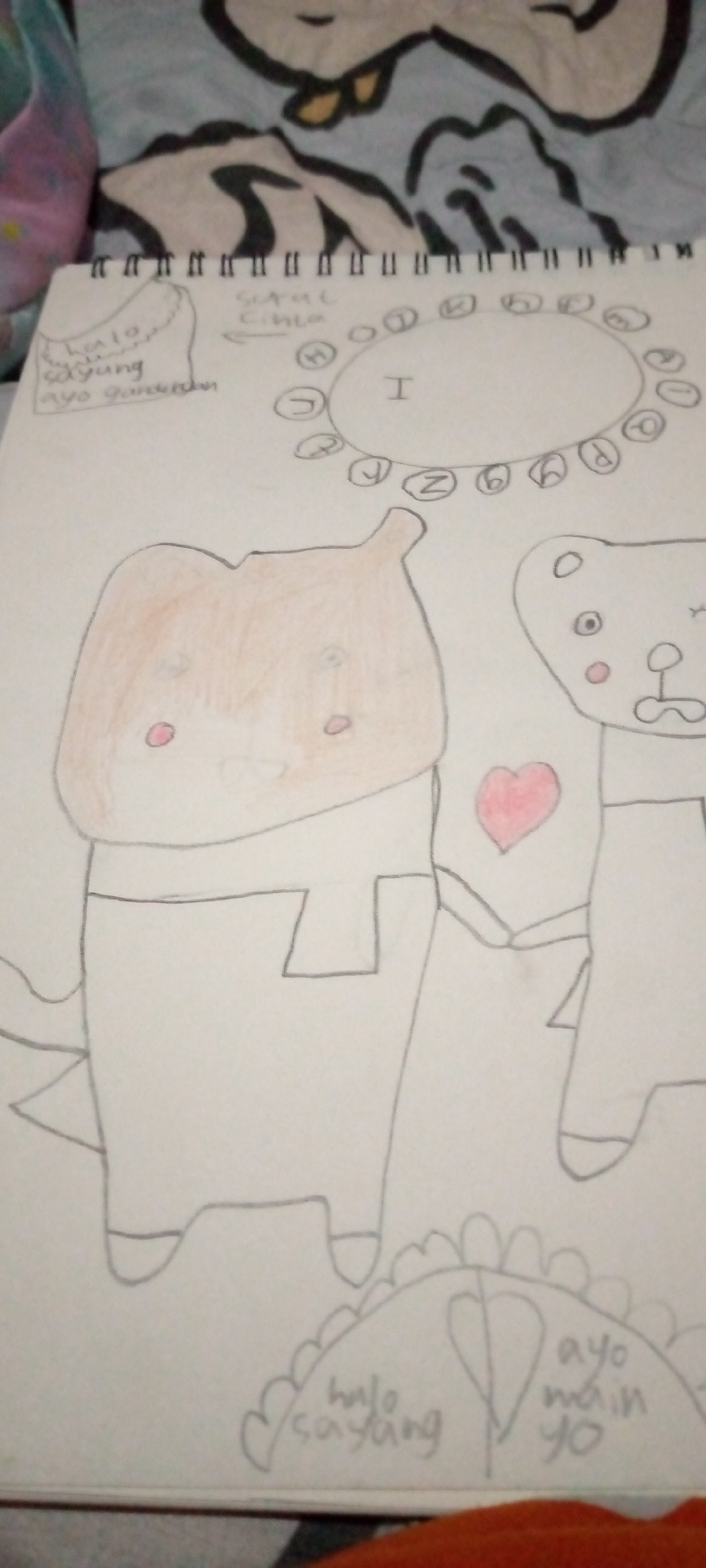 The image shows a hand-drawn illustration on a notebook, with a patterned fabric in the background. The illustration features two cartoonish characters, one appearing to be a brown-colored dinosaur-like creature with round eyes and rosy cheeks, and the other a white teddy bear with similar features, including rosy cheeks and a red heart on its chest. Above the characters, a circular arrangement of letters encircles a central oval containing the letter "I". To the left of the illustration, there is a text box with handwritten words. Below the characters, there is a semicircular shape with more handwritten text. The notebook is open, revealing a spiral binding. The overall scene is domestic, likely a child's drawing. There is no discernible connection to the city of Sumberwono, Indonesia.