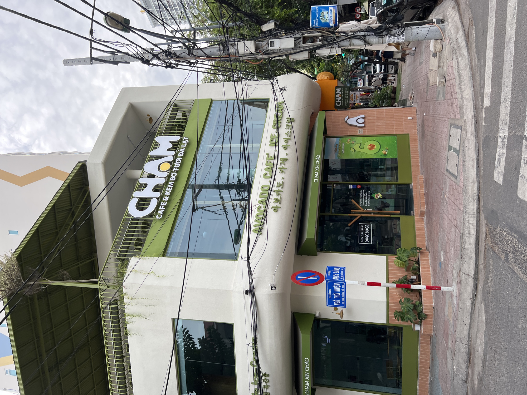The image captures a street-level view of the CHAM coffee and ice cream shop in Nha Trang, Vietnam. The building features a white facade with prominent green accents, including a large awning over the second floor and framing around the windows. The shop's name, "CHAM," is displayed in large white lettering on the front of the building, along with the Vietnamese phrase "CAFÉ & KEM BỞ TƯƠI ĐÀ LẠT" and Korean text "아보카도. 카페. 아이스크림."
A busy intersection is evident with a complex network of electrical wires overhead, typical of urban Vietnam. A traffic light is visible in the distance, indicating a junction. On the sidewalk in front of the shop, a red and white striped pole stands, adorned with street signs in Vietnamese. One sign indicates "ĐƯỜNG NGÔ THỜI NHIỆM" and another "ĐƯỜNG BẠCH ĐẰNG". A blue and white no-stopping sign is also mounted on the pole.
Several potted plants, including large monstera plants, are placed along the building's base. Two people are walking on the sidewalk in front of the shop, and cars are visible further down the street. The shop's entrance area is visible with green framed windows displaying the interior. A sign on the window indicates opening hours from 7:30 to 22:30. The ground is paved with brick-like tiles in front of the shop, transitioning to concrete sidewalk and a paved road with a crosswalk. The sky is overcast.