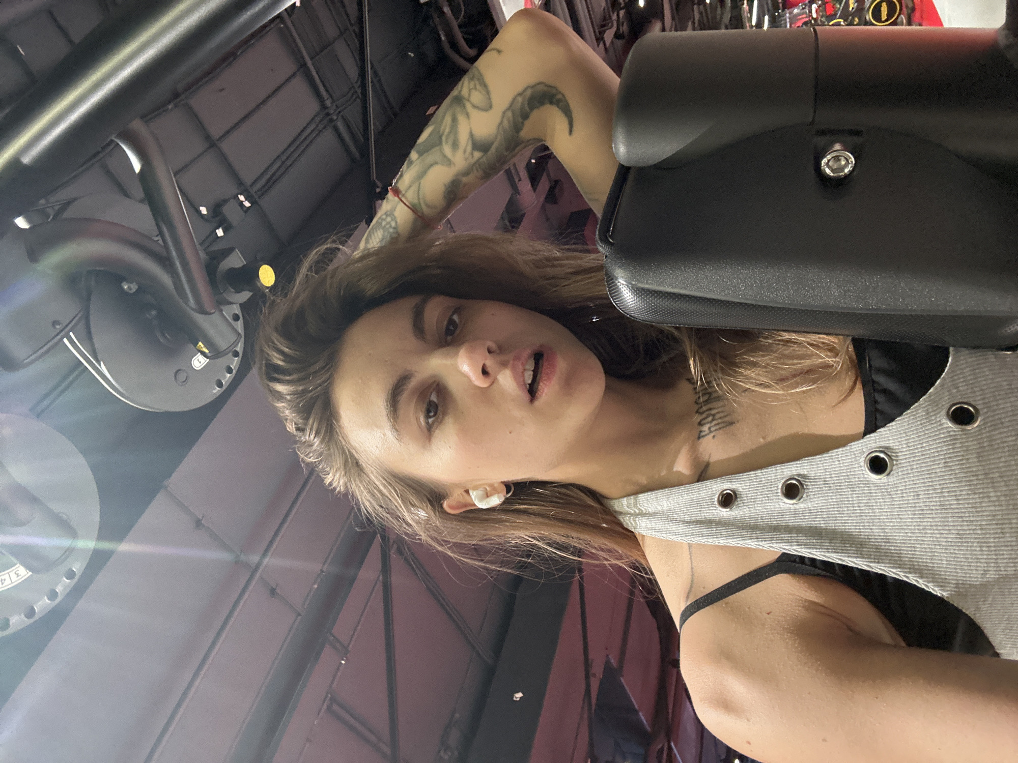 A young woman with long, wavy brown hair is looking up at the camera, her mouth slightly open. She is wearing white earbuds and a grey ribbed halter top over a black tank top. She has a tattoo on her left arm and a red bracelet on her right wrist. She appears to be in a gym, with gym equipment visible in the background. The lighting suggests it is indoors.