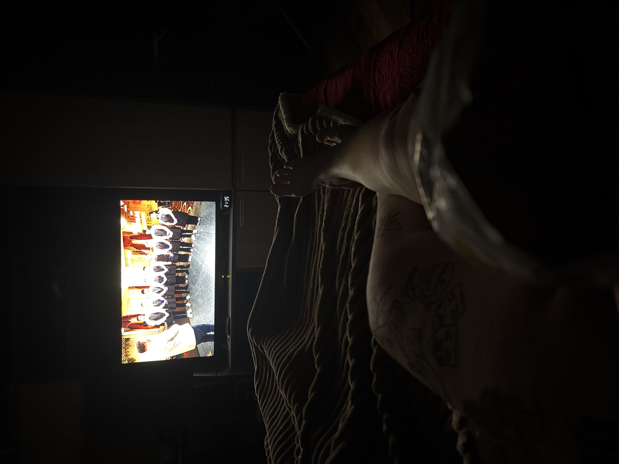 A dimly lit interior view features a television screen displaying a group of individuals in formal attire, possibly a graduation ceremony, given their robes and hats. The scene is captured at an angle, with the focus on the foreground elements. A person's arm and hand holding what appears to be a glass are partially visible, and a tattoo can be seen on the arm. A textured, possibly knitted or rope-like fabric is draped nearby. The background is dark and mostly obscured. The location in the city of Chornomorskoe, Ukraine is not discernible from the visual information provided.