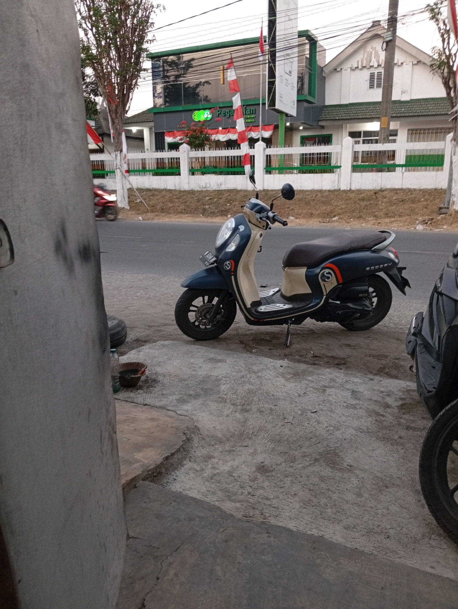 A dark blue and beige scooter is parked on a concrete surface next to a road. The scooter is facing right and is the primary subject of the image. Behind the scooter is a white fence with green accents, and beyond that, a building with a green-tinted glass facade and a white structure with a green roof. The building appears to be an office or commercial establishment. Indonesian flags are visible on poles in front of the building. The ground is covered in cracked concrete and dirt. A large concrete pillar is in the foreground on the left side of the image. The lighting suggests it is either early morning or late afternoon.