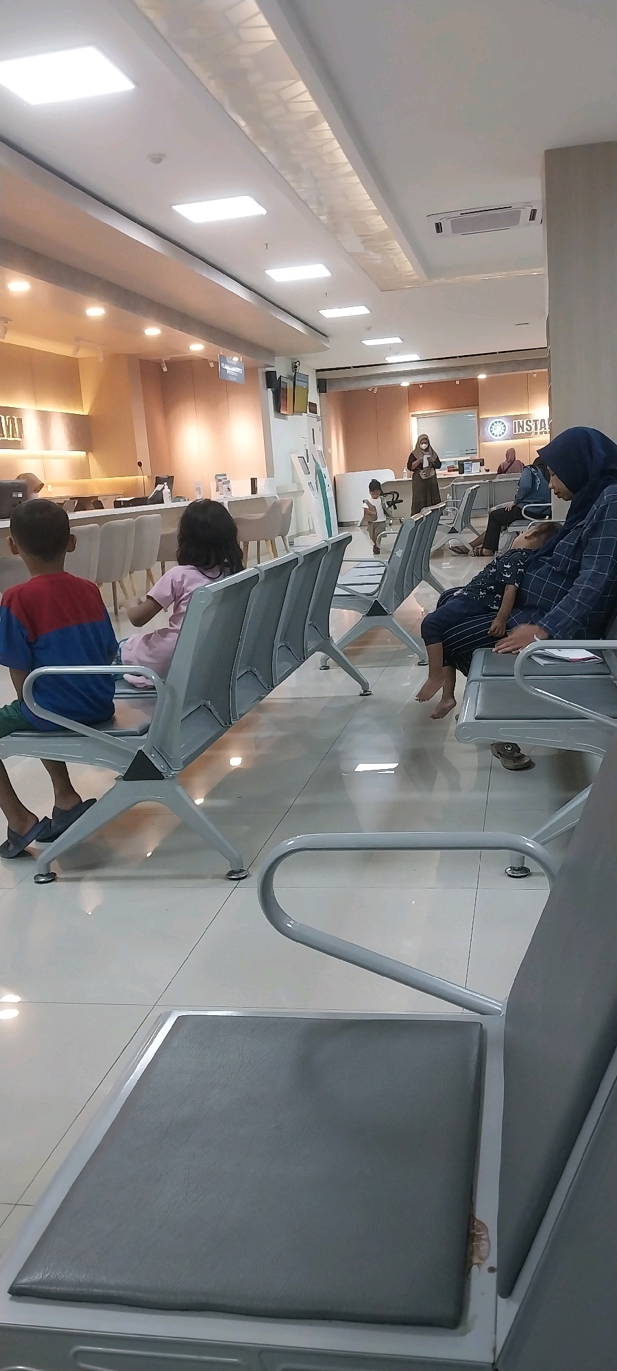 The image depicts the waiting area of a clinic in Kebonagung, Indonesia. Several people are seated on a row of grey metal chairs arranged in a waiting room. In the foreground, two children are visible, one facing away from the camera and the other from behind. Further back, a woman wearing a blue hijab is holding a sleeping child on her lap. In the background, a woman in a brown headscarf stands near a reception desk, possibly interacting with patients. The environment is brightly lit by overhead fluorescent lights and appears clean with tiled flooring. The clinic's interior design suggests a modern and functional space.