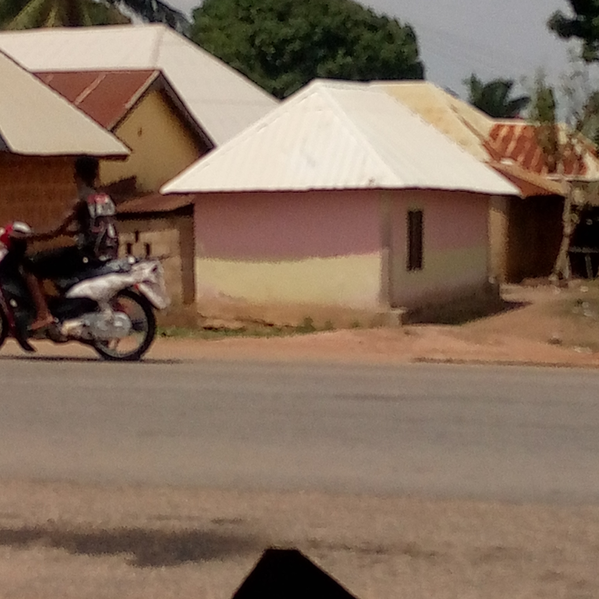 The image depicts a street scene in a location identified as Unknown, Unknown. In the foreground, a paved road stretches across the frame. A person rides a white motorcycle on the left side of the road. In the background, there are several buildings of varying styles and conditions, including a pink and yellow building with a white roof, as well as buildings with red roofs. Trees and vegetation are visible between and behind the buildings. The sky appears overcast or hazy.