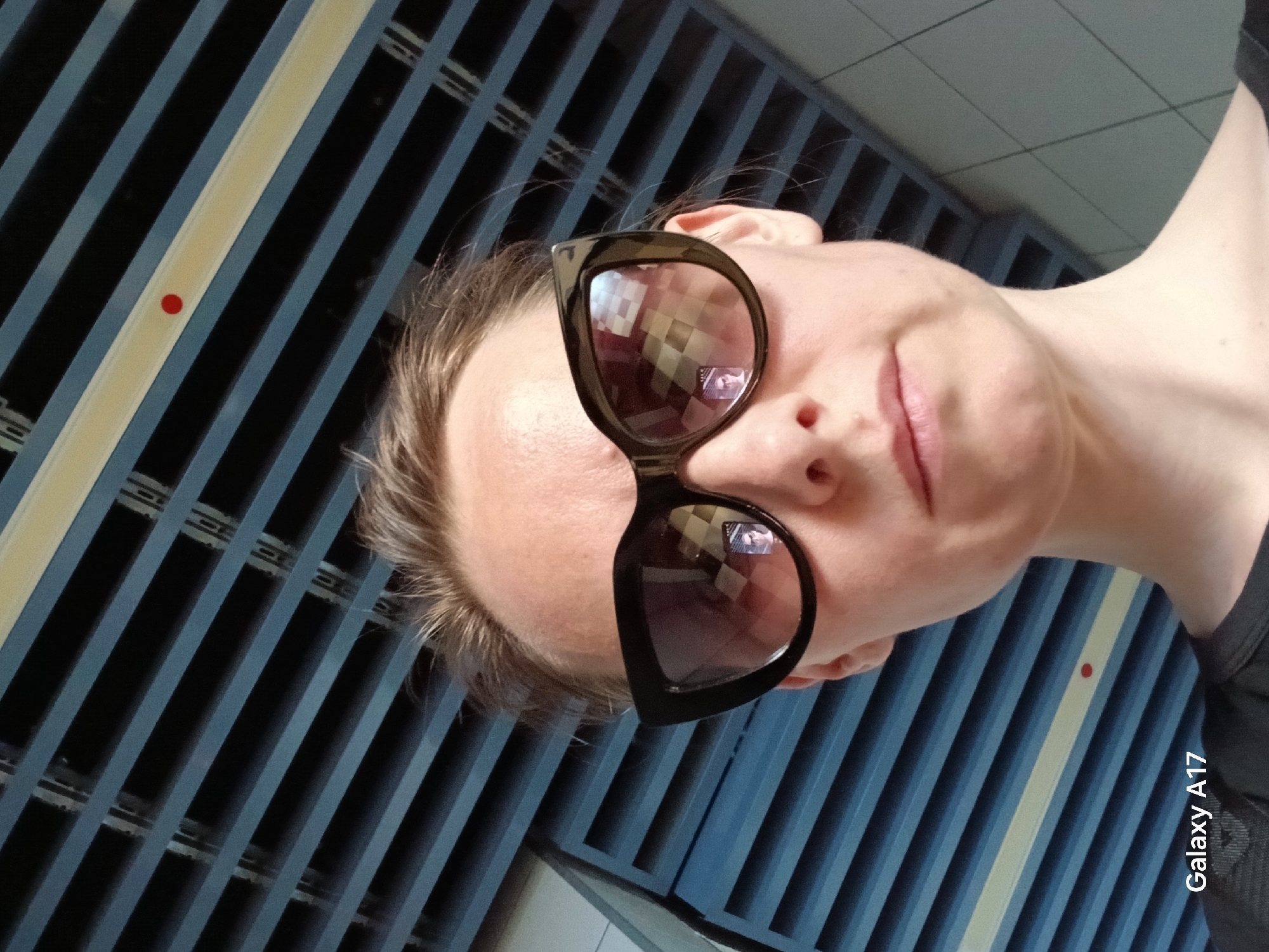 A woman with fair skin is wearing large, black sunglasses and is positioned in front of a background of blue and white striped metal bars, possibly a fence or gate. The reflection in her sunglasses shows a checkered or grid-like pattern, suggesting an urban environment. The photograph appears to have been taken in Yekaterinburg, Russia, based on information embedded in the image file (likely geotagged metadata) which is now illegible. The woman’s face is partially visible, showing her nose, lips, and part of her cheek. The angle suggests a selfie perspective. "Galaxy A17" is visible on the bottom right corner, indicating the device used to take the picture.