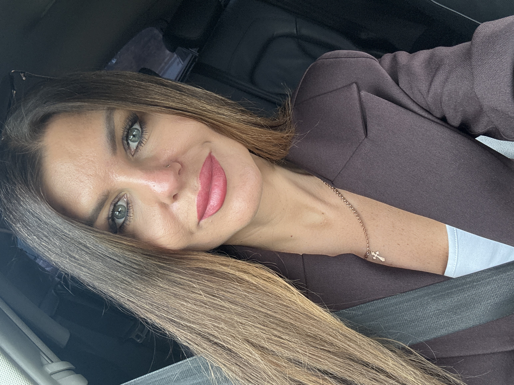A woman with long, brown hair and blue eyes, wearing a dark purple blazer, is seen from the chest up, seated in a car. She has bright pink lipstick on and a small cross necklace. The image was taken in the city of Chaikovsky, Russia.