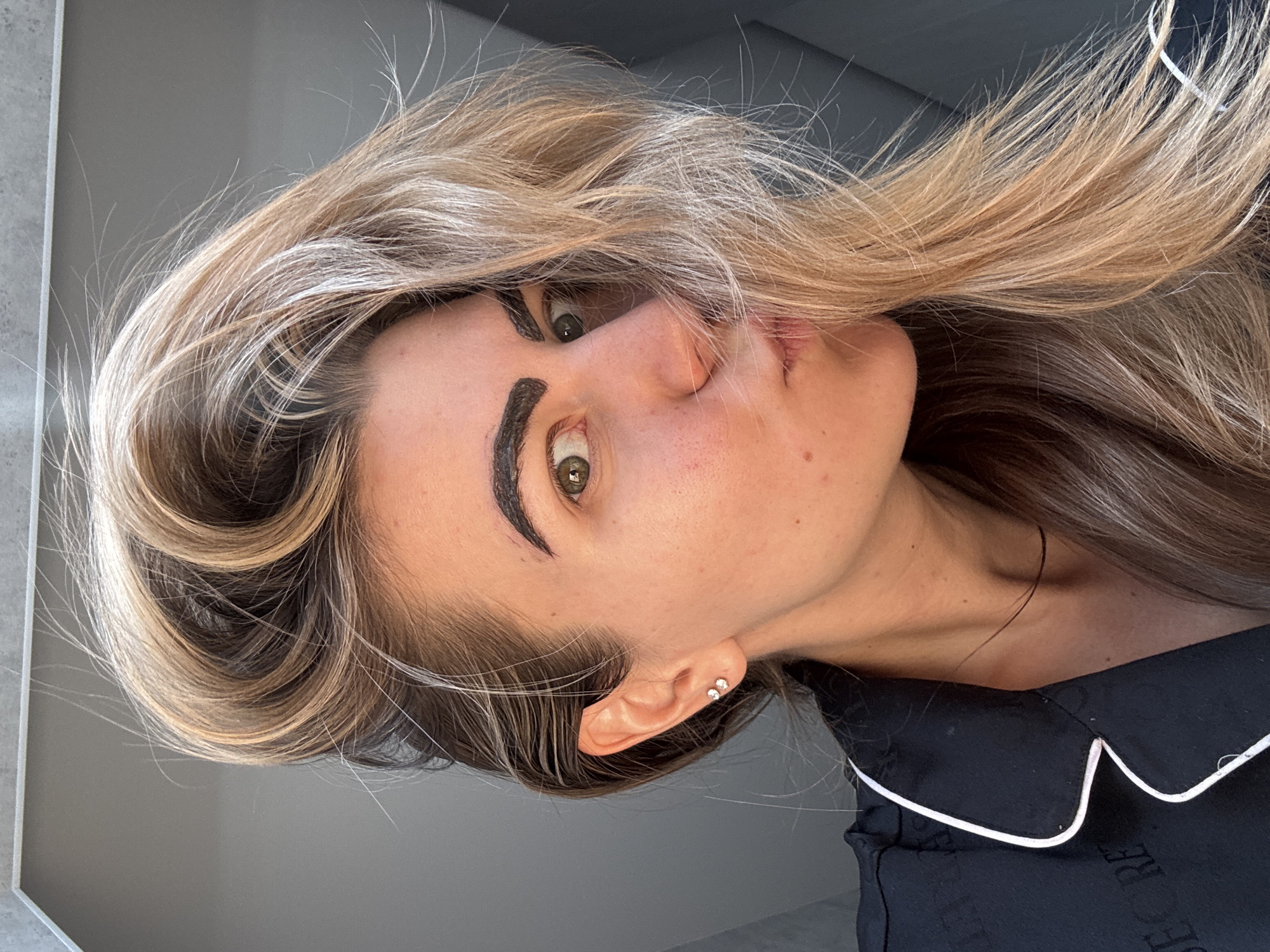 A woman is depicted in a close-up, angled portrait. She has fair skin, light brown and blonde hair styled with volume, and green eyes. Her eyebrows are notably dark and thick. She is wearing a black garment with white piping around the collar. She has two earrings in her ear. The background appears to be a neutral gray wall. The location is Unknown, Unknown.