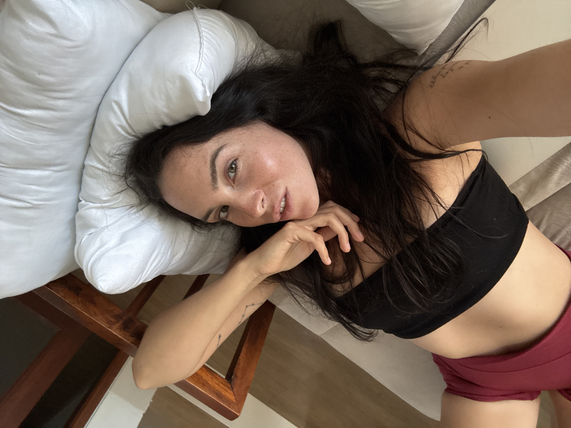 A woman is lying on her back on a couch, propped up by a white pillow, in what appears to be a living room in North Kuta, Indonesia. She has long, dark hair and is wearing a black bandeau top and burgundy shorts. Her left arm is bent and resting near her head, with her hand touching her face. A wooden chair is visible to the left of the couch. The overall lighting suggests it is daytime.