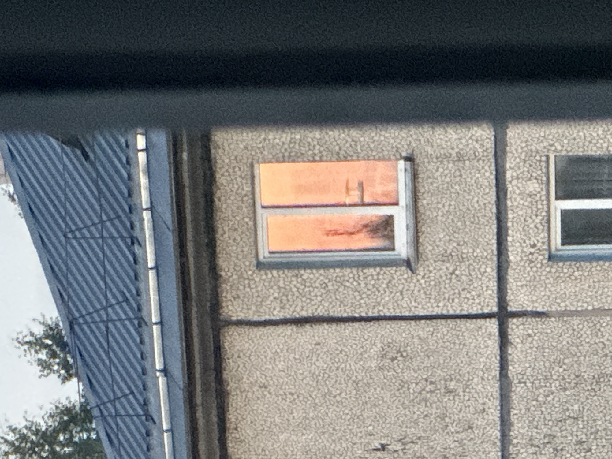 The image shows a close-up view of a building's exterior in Plesetsk, Russia. The building has a textured, light-colored facade and a dark, sloped roof with visible metal sheeting. A window on the upper floor is illuminated with a warm, orange light, reflecting what appears to be a sunset or artificial lighting. The reflection in the window is abstract, showing vertical shapes and hints of what might be trees or structures. The window frame is white. The overall scene is framed by a dark, out-of-focus vertical element on the right side of the image, likely a part of another building or structure.