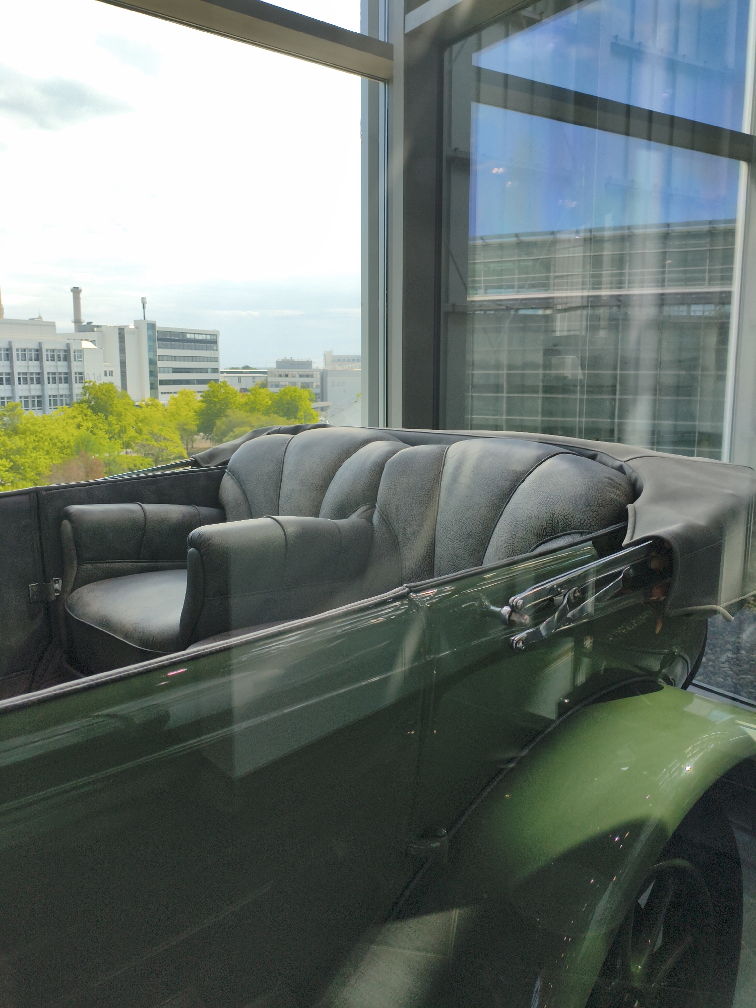 The image shows an interior view of a vintage dark green convertible car. The car has plush leather seats with a tufted backrest and armrests. Through the car's windows, a cityscape is visible, featuring modern buildings and trees. The car appears to be displayed indoors, possibly in a museum, given the reflective glass partitions. The setting is likely a public space within Ingolstadt, Germany, as indicated by the presence of contemporary architecture. No people are present in the image.