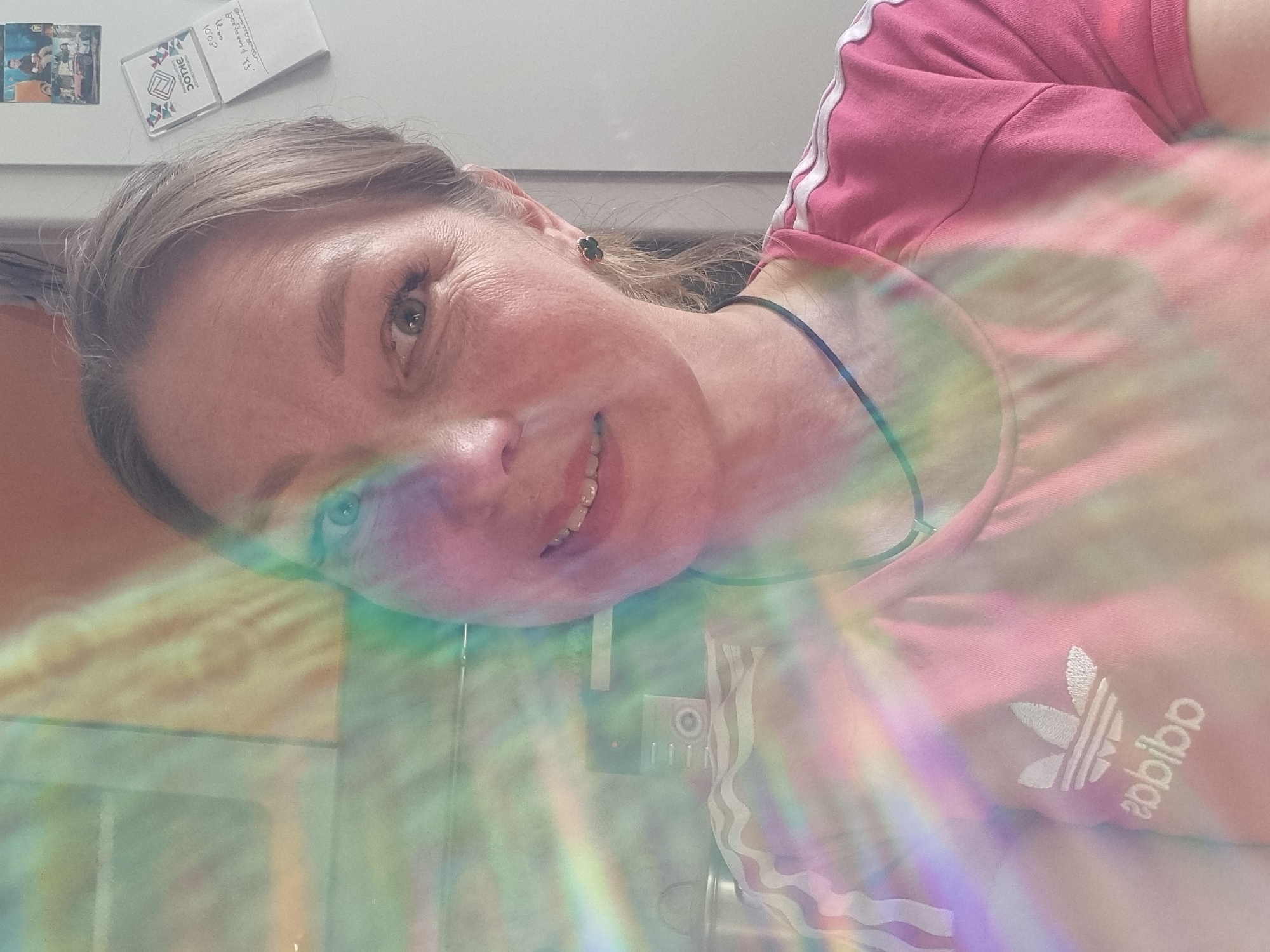 A selfie of a woman wearing a pink Adidas t-shirt, likely indoors. The image includes a kitchen or similar room setting, with appliances and accessories visible. A rainbow light effect is overlaid on the scene. The presence of a refrigerator magnet with Cyrillic text may suggest a location in a Russian-speaking area, possibly in the city of Chaikovsky.