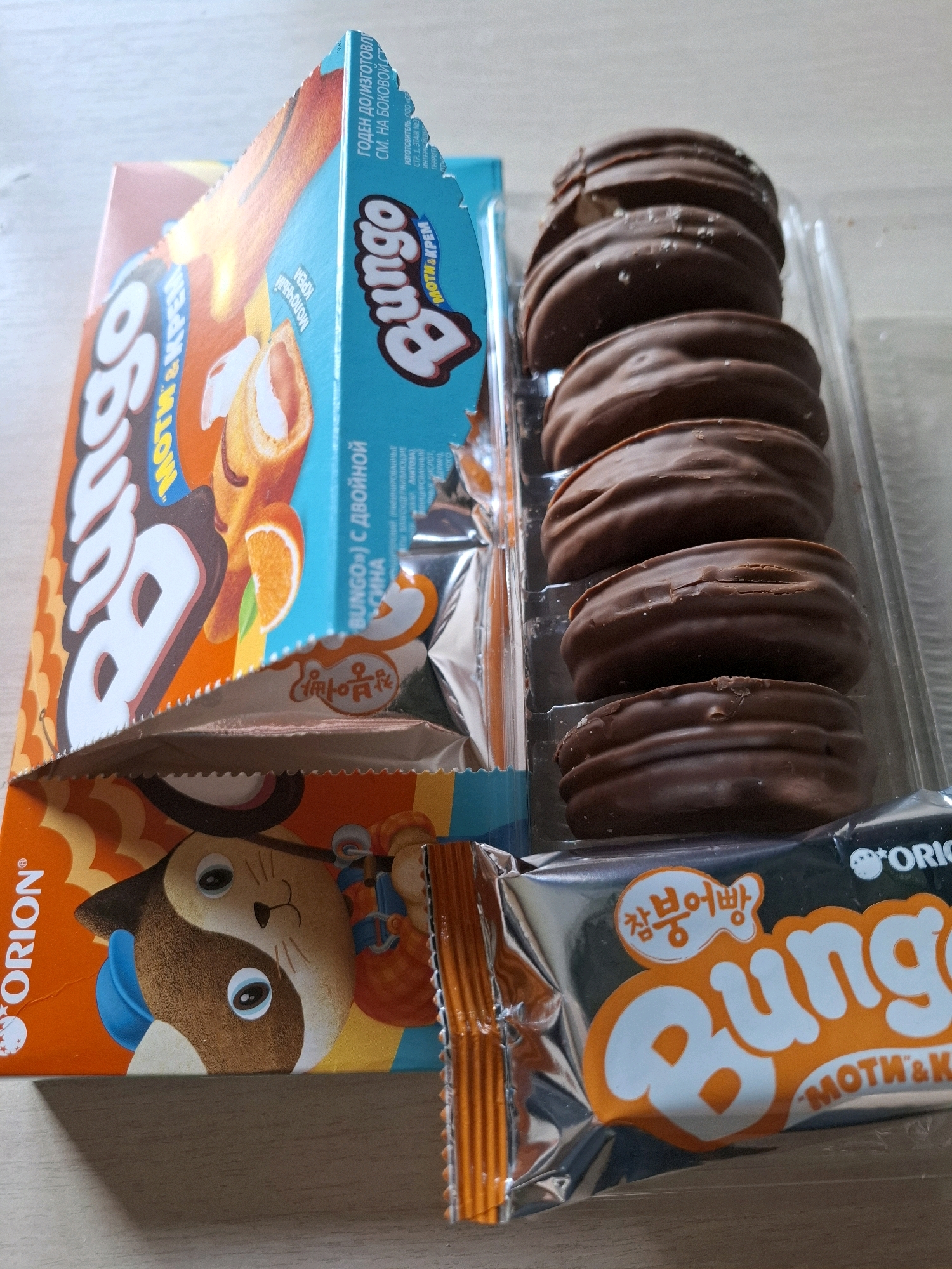 In the city of Tyumen, Russia, a still life image captures a collection of Orion's Choco Pie, also known as "Bungo" cookies. The cookies, coated in chocolate, are neatly arranged in a clear plastic tray, with a slightly open box of the same product visible to the left. The packaging features vibrant orange and blue hues, with the "Bungo" logo prominently displayed in white lettering against an orange background. Some of the cookies have a light dusting of what appears to be powdered sugar or salt. The overall setting suggests a domestic environment, possibly a kitchen counter or table, with a hint of wooden texture in the background. The lighting is soft and diffused, creating gentle shadows.