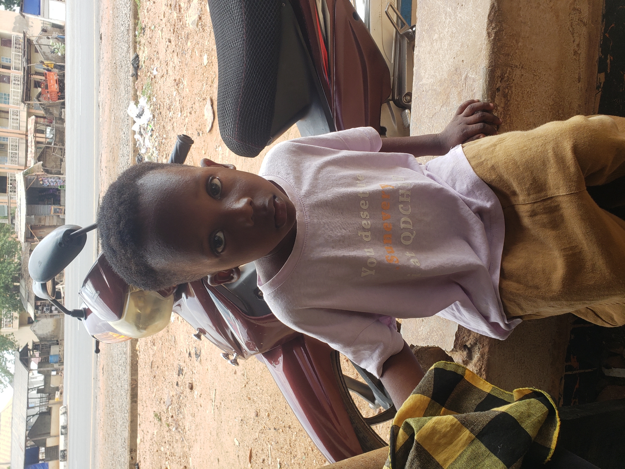 A child sits on a maroon motorcycle parked on a dusty surface in Adankolo, Nigeria. The child wears a pale purple t-shirt and khaki pants. The motorcycle has a black seat, a yellowed headlight, and side mirrors. In the background is a road and various buildings. A checkered yellow and black cloth sits near the child's legs. The child is looking directly at the camera. The picture has been rotated.