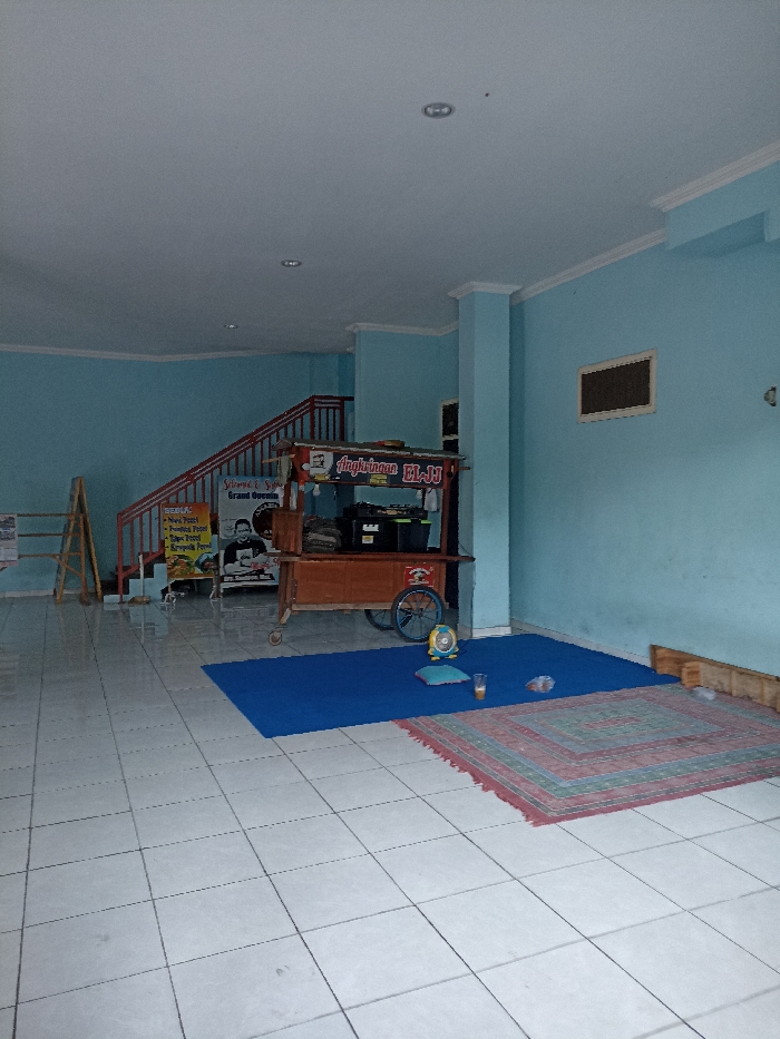The image depicts an empty indoor space, likely a casual eatery or gathering spot, with light blue walls and a white tiled floor, characteristic of a commercial area in Tulungagung, Indonesia.
A prominent wooden food cart, identified by the text "Angkringan EL-JJ," stands in the middle ground. To the cart's left, a promotional banner for a "Grand Opening" is visible, featuring the phrases "Selamat & Sukses" and "BERKAH," alongside a list of Indonesian food items such as "Nasi Kucing," "Sate Usus," "Sate Telur," "Sate Bakso," "Sate Jamur," "Sate Kulit," "Pindang Pete," "Pindang Kerupuk," "Telpo Kerupuk," and "Kerupuk Pecel." A picture of a man is also on the banner.
Further to the left, a wooden staircase with red railings leads upwards. A wooden A-frame ladder leans against the wall near the staircase. On the floor to the right of the food cart, a large blue mat is laid out, partially covered by a smaller, patterned rug in shades of red, blue, and green. On the blue mat, there's a small light blue pillow, a glass containing a drink, and a compact yellow fan. The ceiling is white with multiple recessed light fixtures, and a small, dark rectangular window is set into the light blue wall on the far right. The space is well-lit, suggesting it is daytime. No individuals are present in the scene, which appears quiet and set up for future activity.