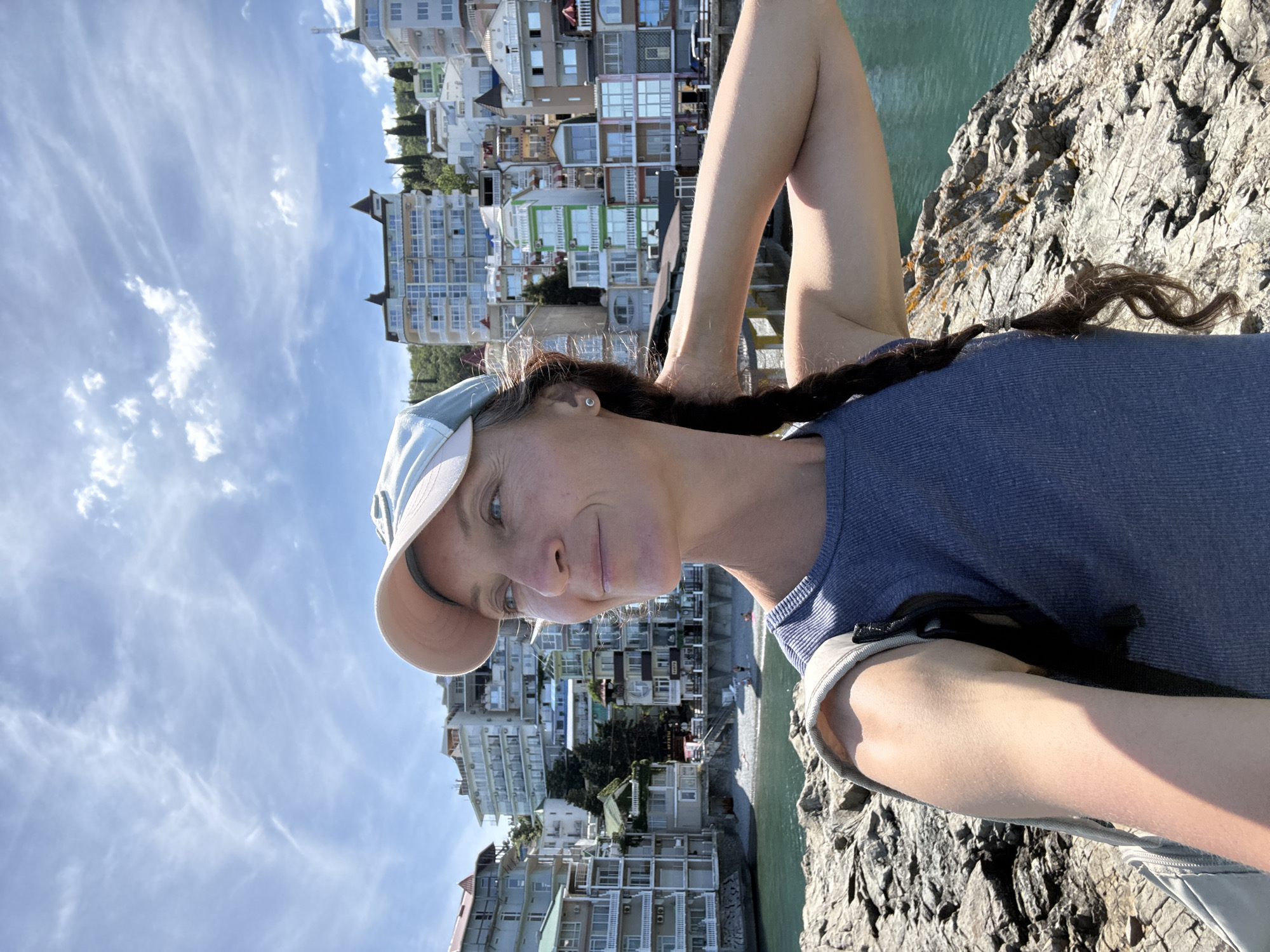 The media file is a portrait-oriented selfie of a woman taken outdoors. She is wearing a blue tank top and a light-colored baseball cap. Her hair is dark brown and styled in a braid that is draped over her shoulder. She is smiling at the camera.
Behind the woman, the scene shows a steep, rocky incline to her right, with the turquoise water of the sea visible in the background. To her left, a densely packed collection of multi-story buildings with balconies and windows is visible, suggesting a coastal town or resort. The buildings are varied in color and architectural style, with some appearing to be hotels or apartment complexes. Trees and greenery are interspersed between the buildings and on the slopes. The sky above is blue with scattered, wispy white clouds.
The lighting suggests it is daytime, likely sunny given the shadows and the brightness of the scene. The woman's expression appears relaxed and happy, perhaps enjoying a vacation or a scenic outing. The overall impression is one of a pleasant day at a coastal location, with the woman capturing a moment of her experience. There is no visible text in the image. The location is contextually identified as Utios, Ukraine.